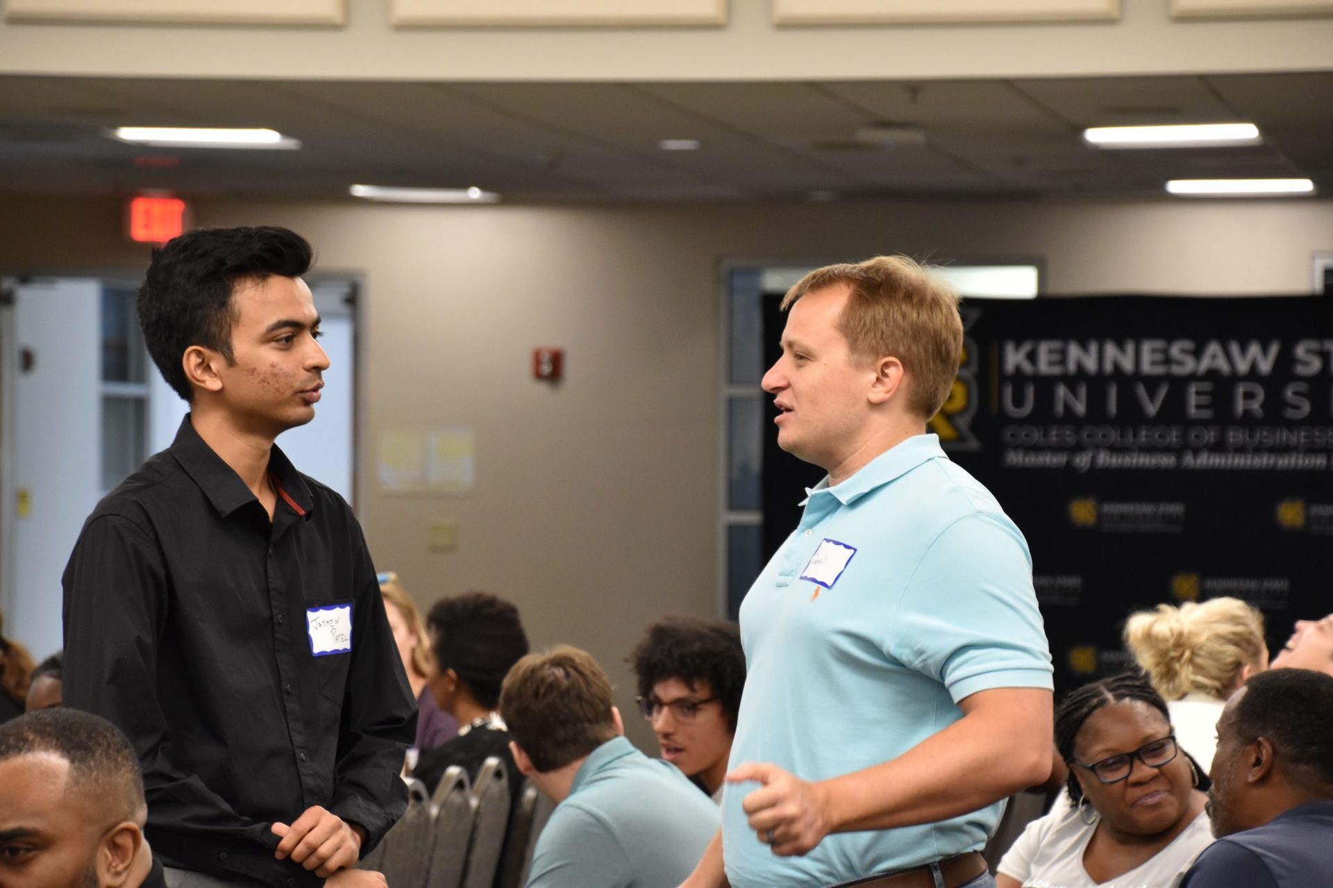 Two men, one in a black shirt and one in a blue polo, talk at a Kennesaw State University event.