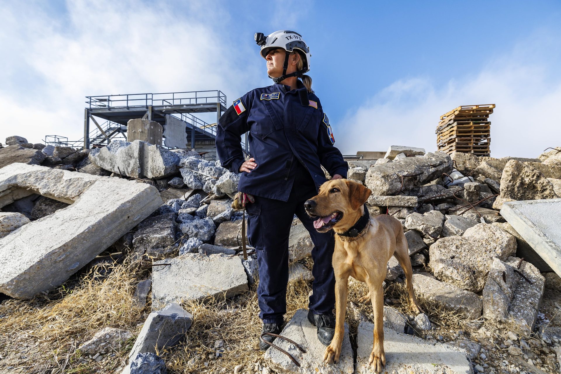 A female search and rescue worker and her dog stand in a debris field.