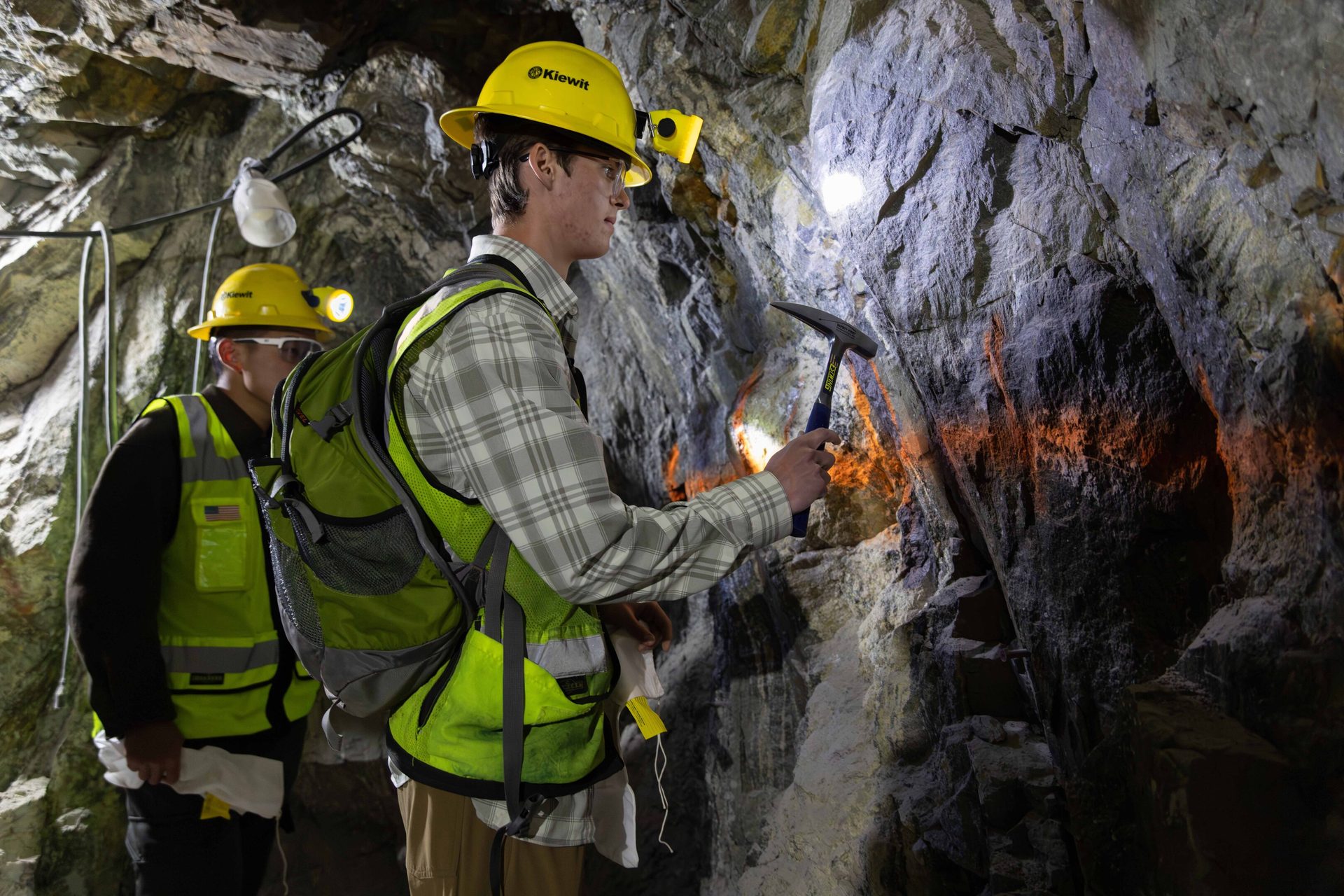 Two people in mining gear inspecting cave walls with a hammer and headlamps.