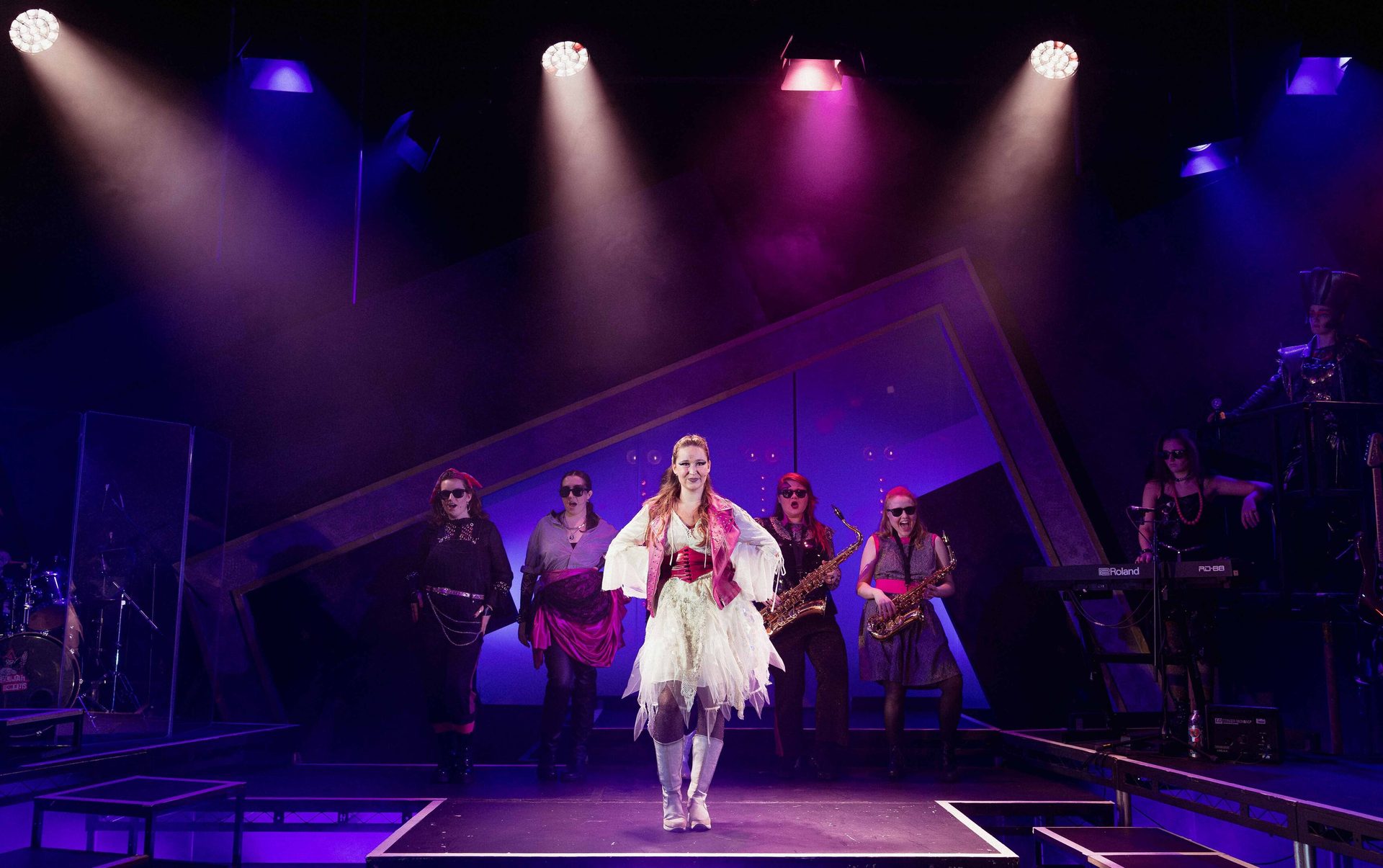A lead singer performs on stage with her band, under bright purple and pink spotlights.