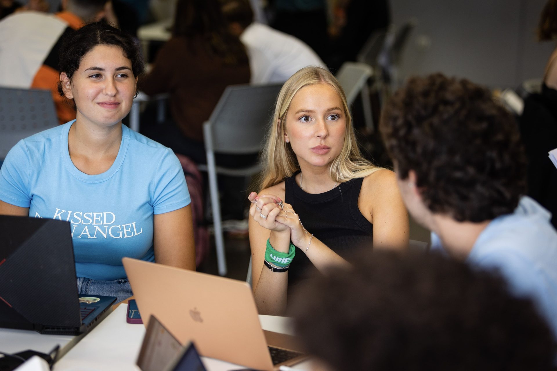 Two young women with laptops, one in a blue shirt, one in a black top, engaged in a discussion.