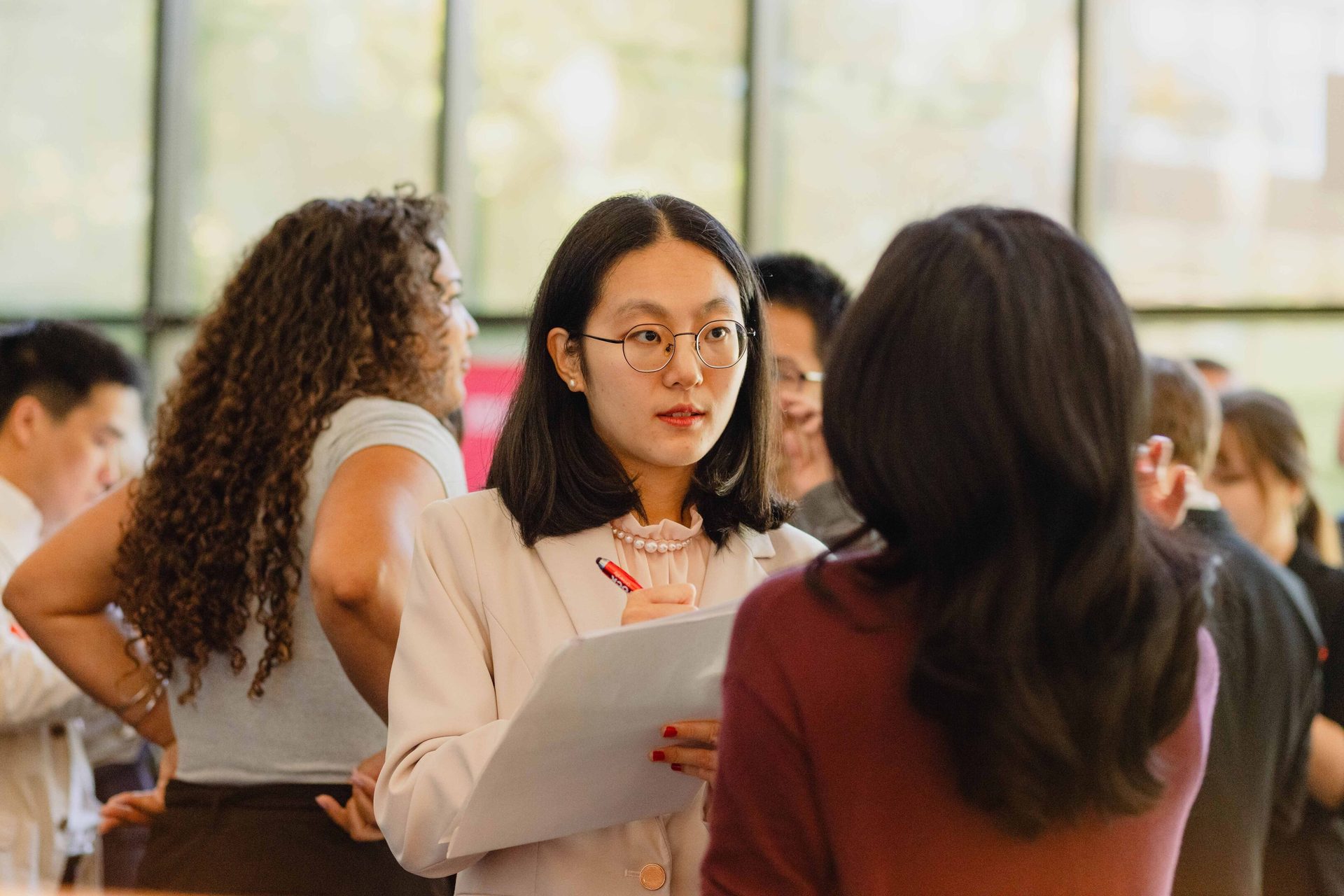 A young woman in glasses and a blazer takes notes while speaking to another person.