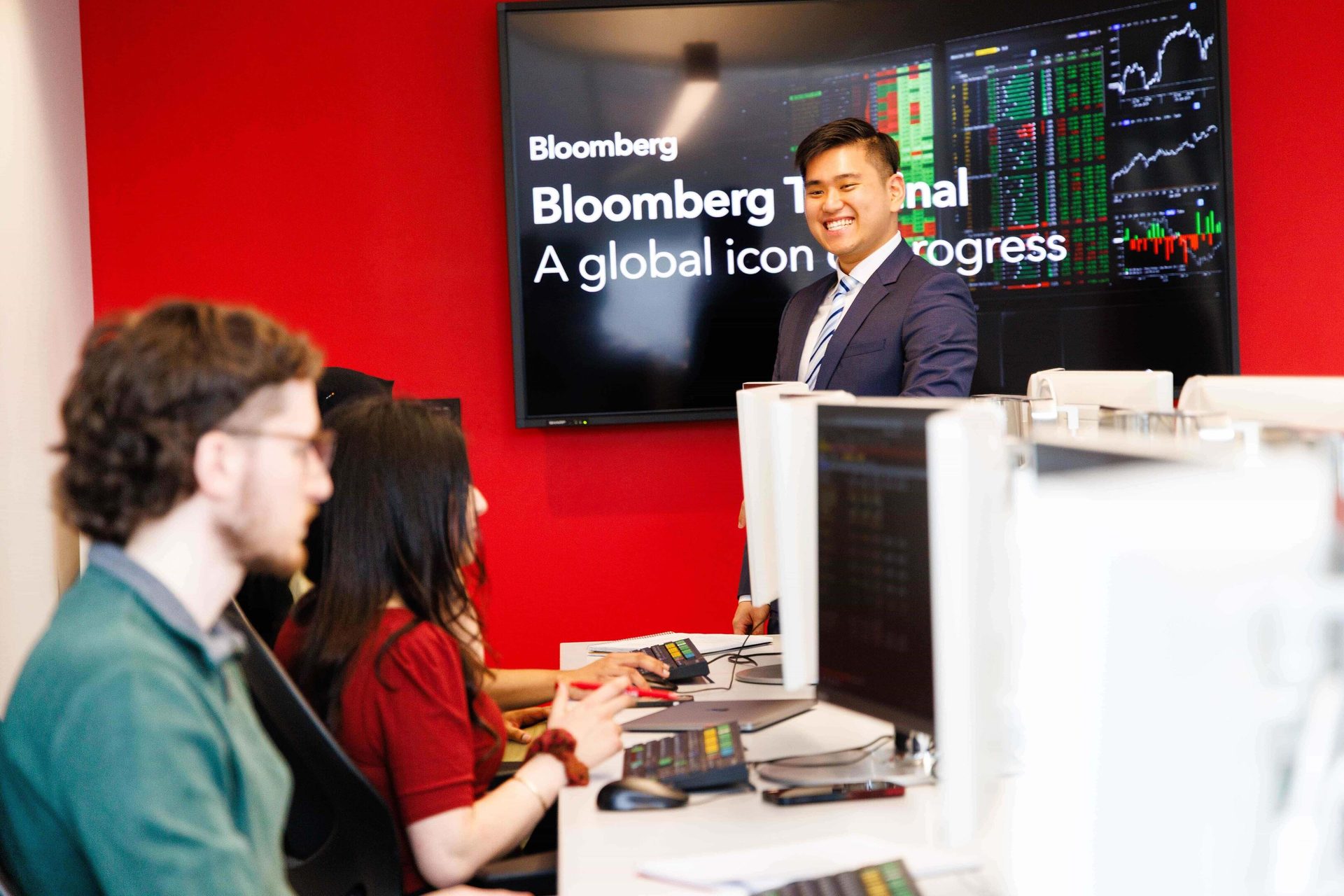 Smiling man in suit by Bloomberg screen with financial data; others sit at desks.