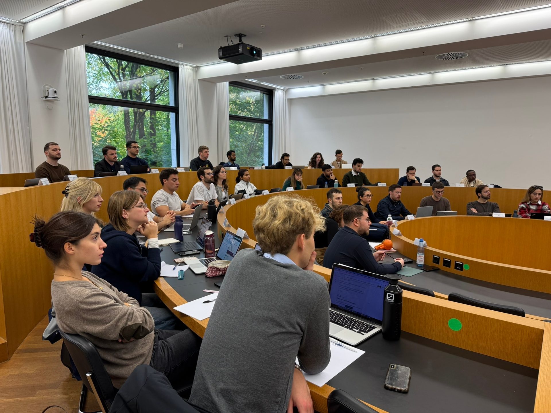 Many students seated in a modern, tiered lecture hall with curved desks, laptops, and large windows.