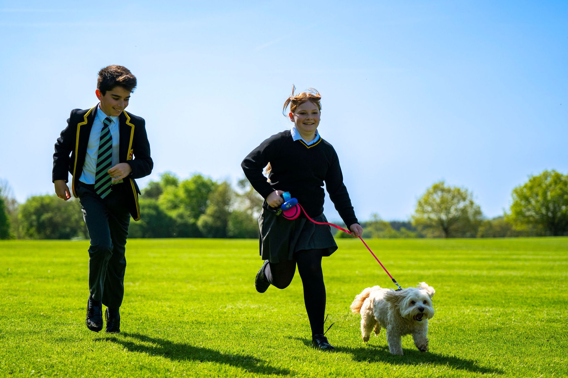 Two school children in uniform and a dog run across a sunny green field, enjoying the outdoors.