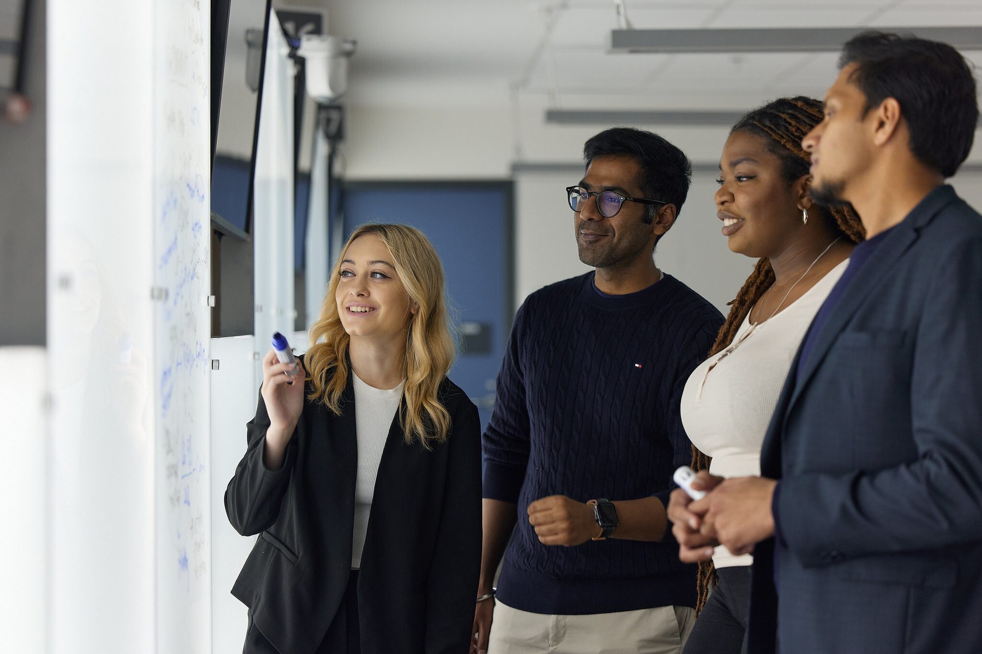 Four diverse professionals collaborate at a whiteboard in an office.