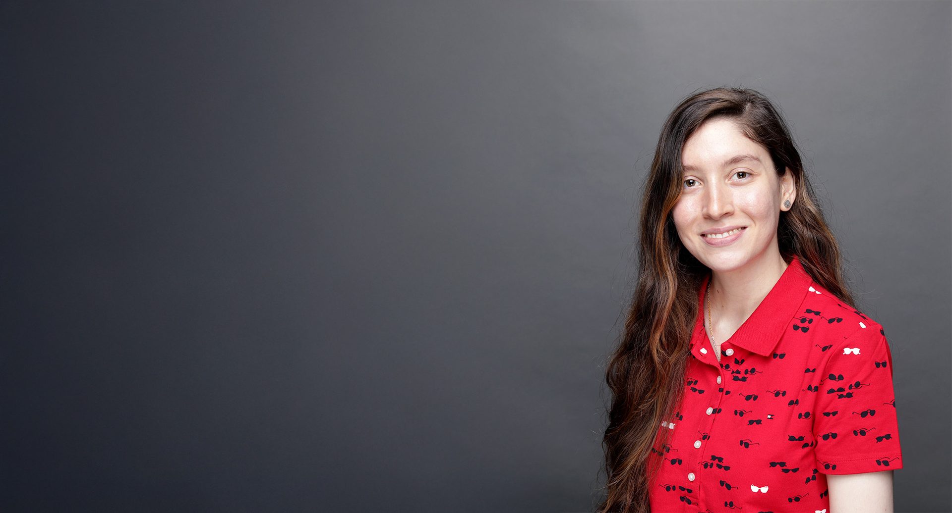 Smiling woman with long hair, wearing a red polo shirt patterned with sunglasses, against a gray background.
