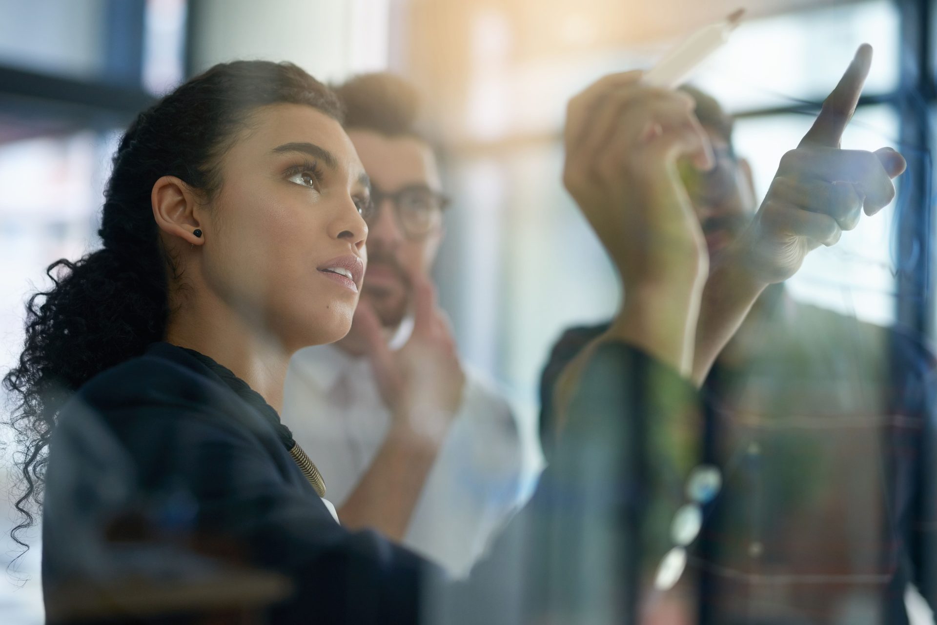 A woman observes intently as two colleagues brainstorm on a clear board in an office.