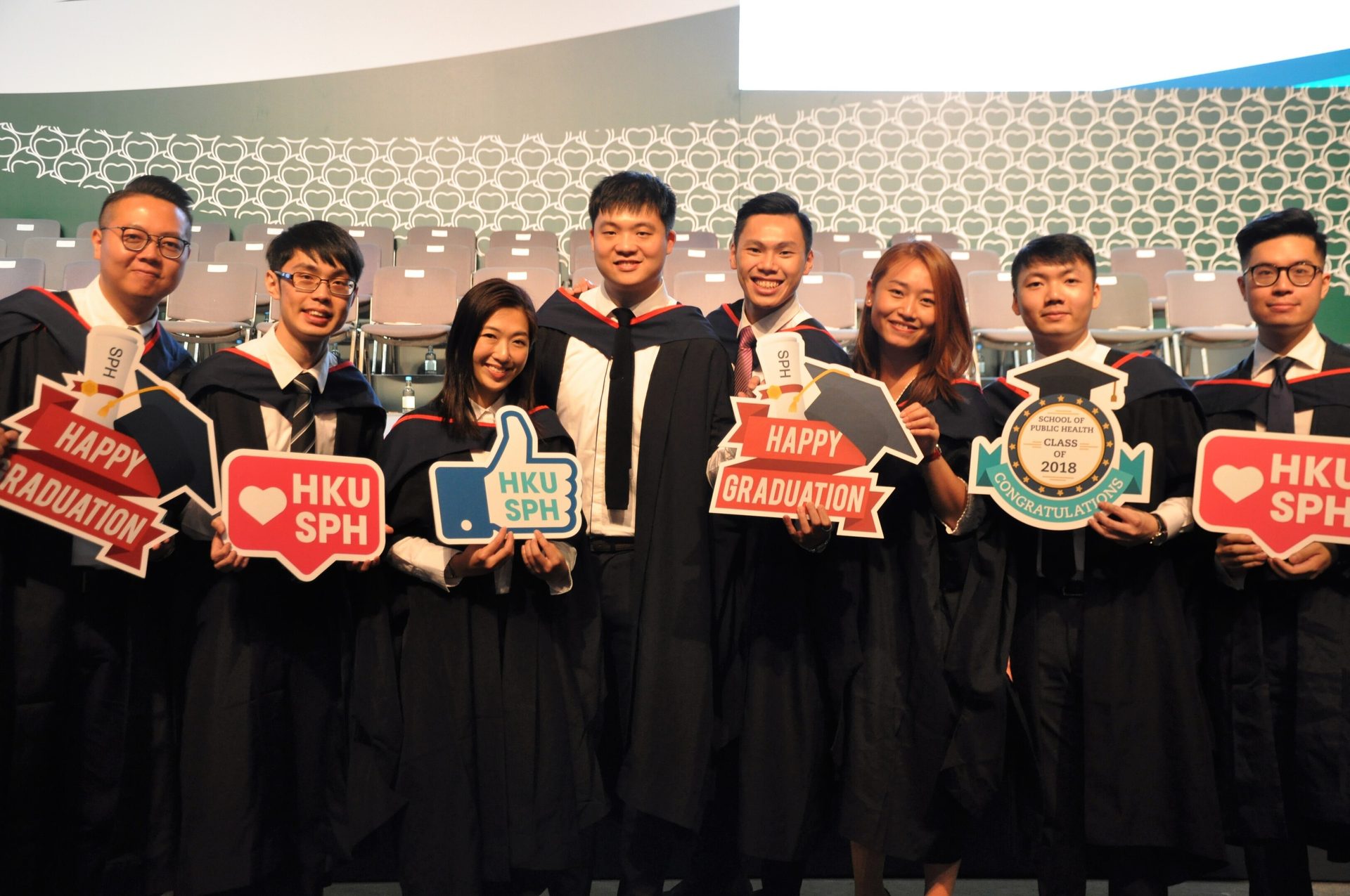 HKU SPH graduates in robes hold "Happy Graduation" & "Class of 2018" signs.