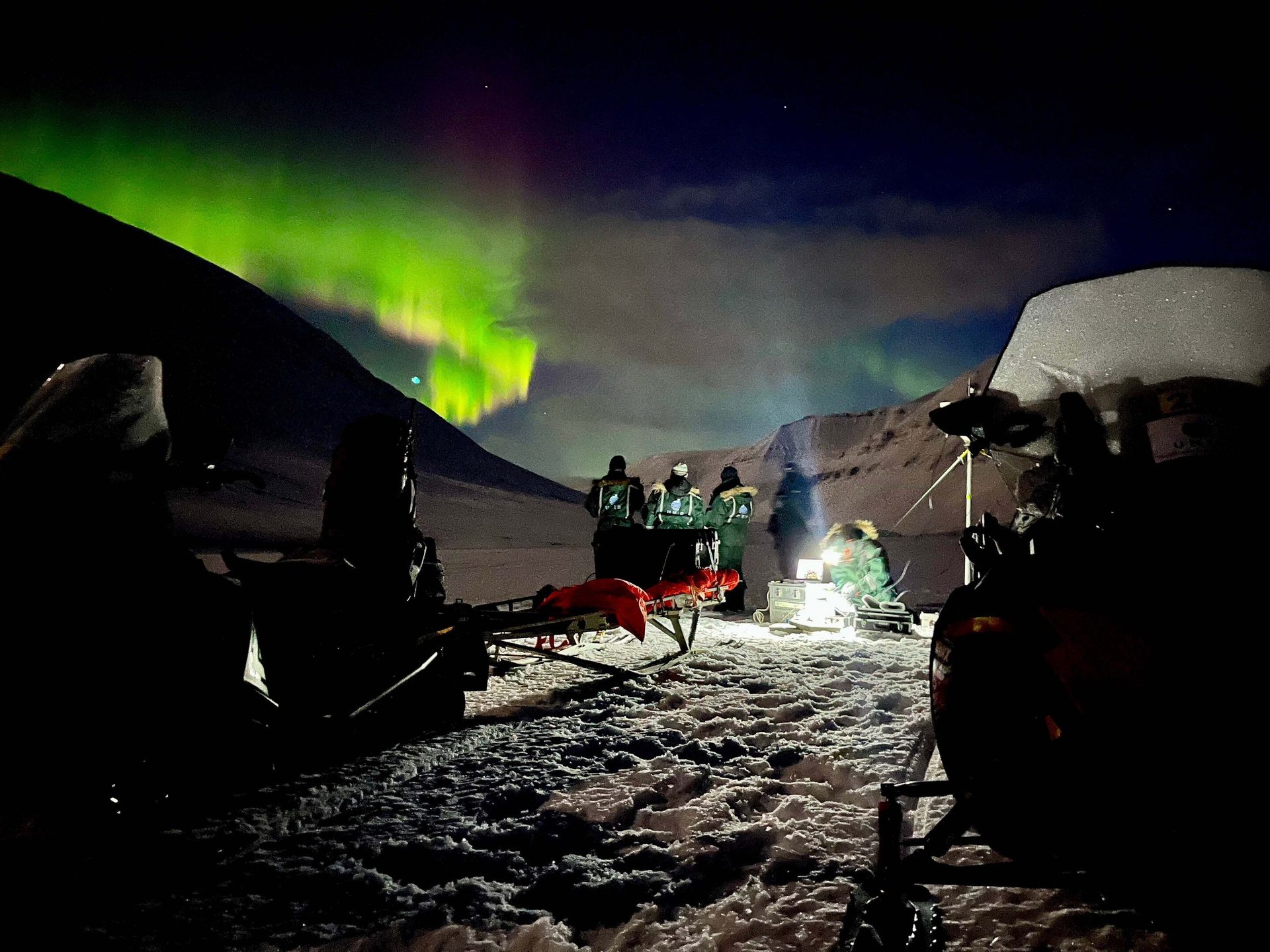 People and equipment in a snowy arctic landscape at night, illuminated by bright green aurora borealis.