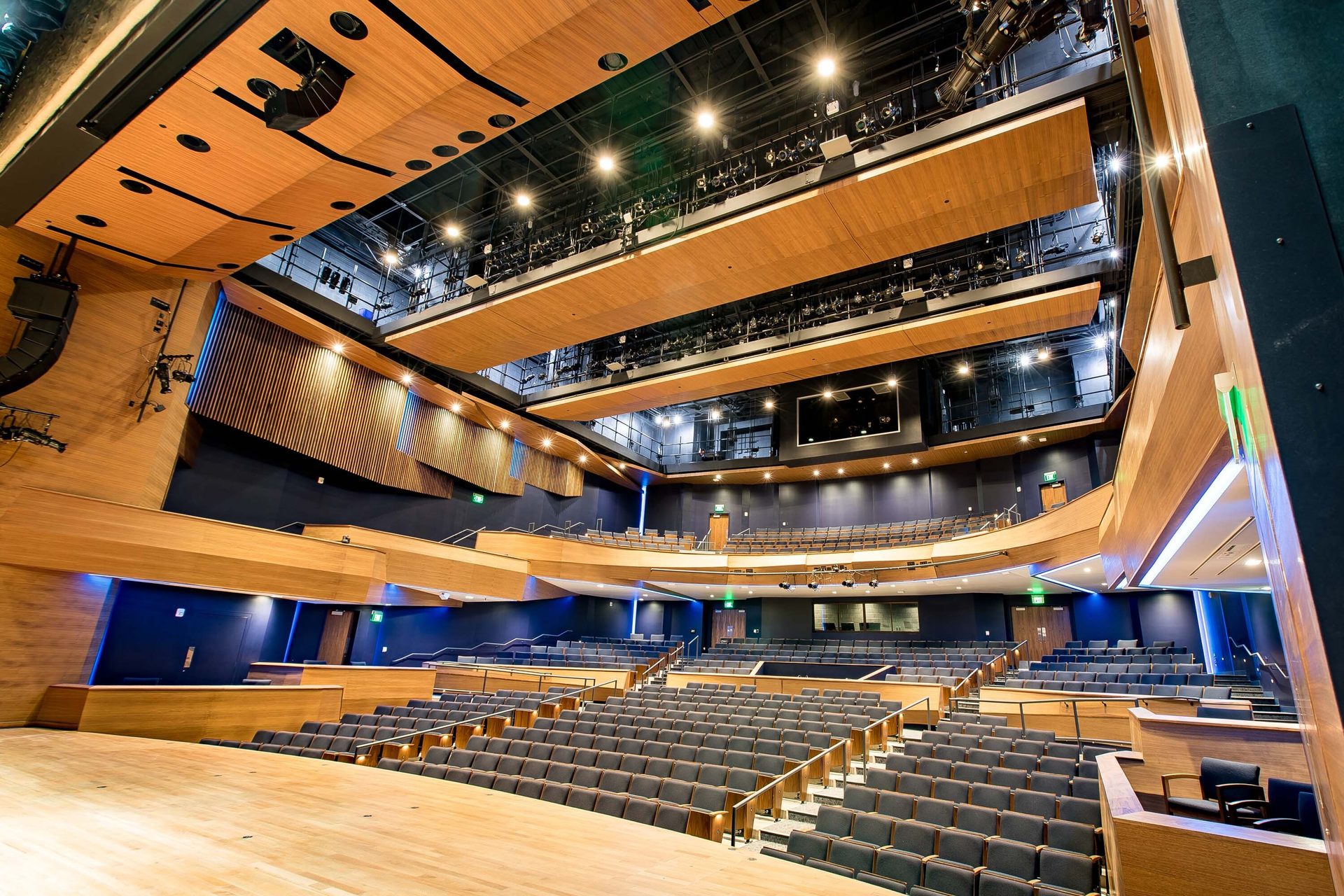 Modern auditorium with light wood panels, dark walls, and rows of blue seats facing a stage.