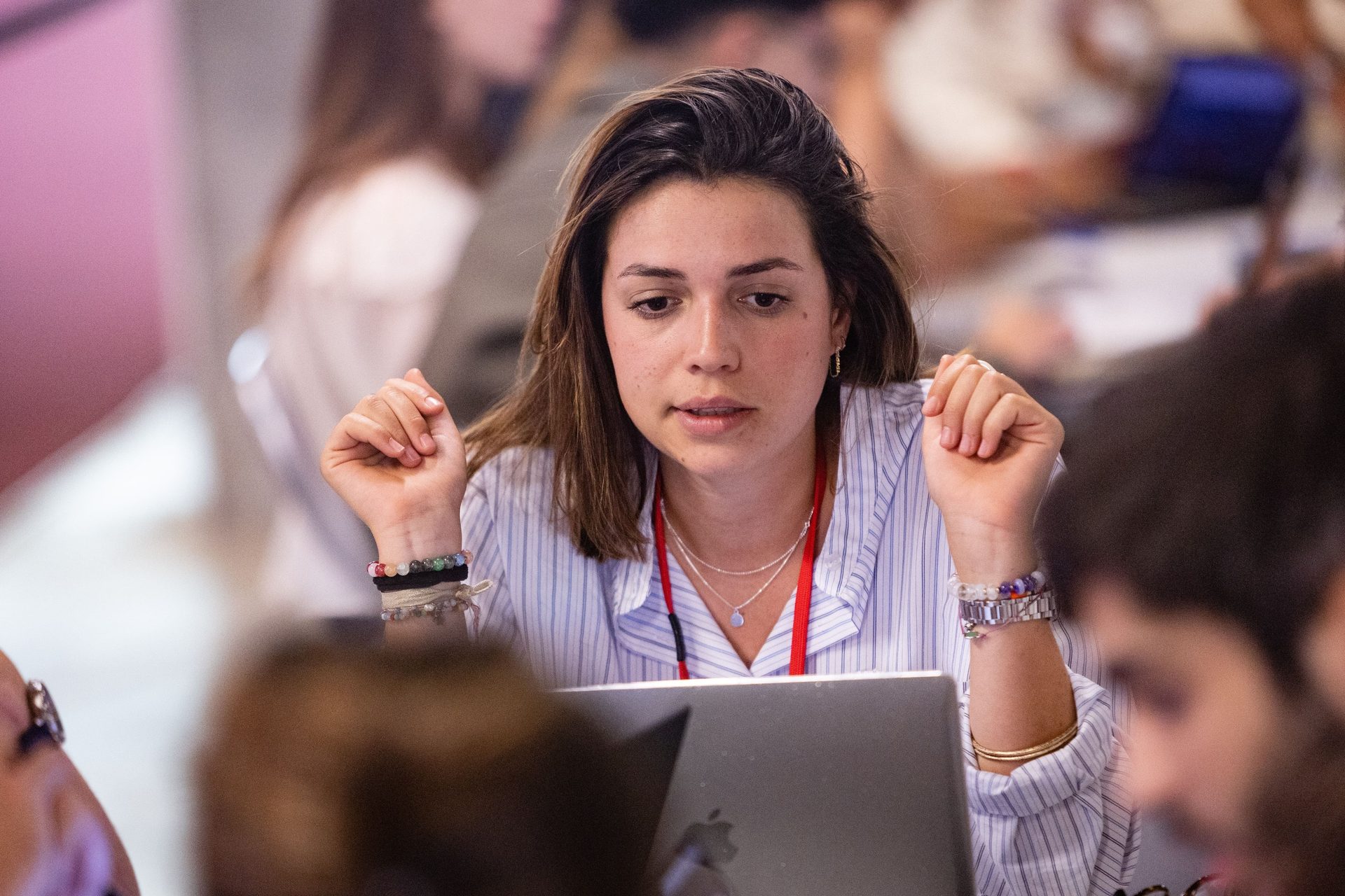 Woman focused on laptop, hands gesturing, in a lively group environment.