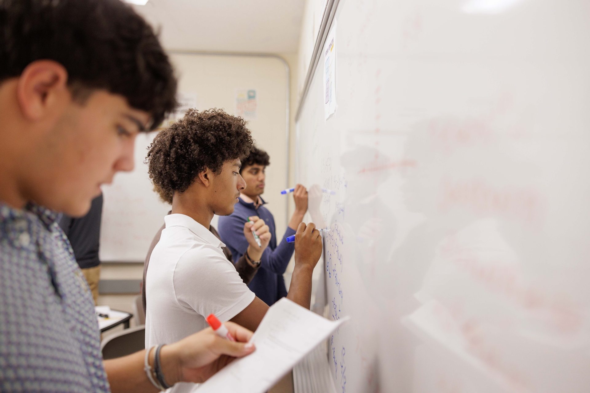 Three male students are actively writing on a whiteboard and reviewing papers in a classroom.