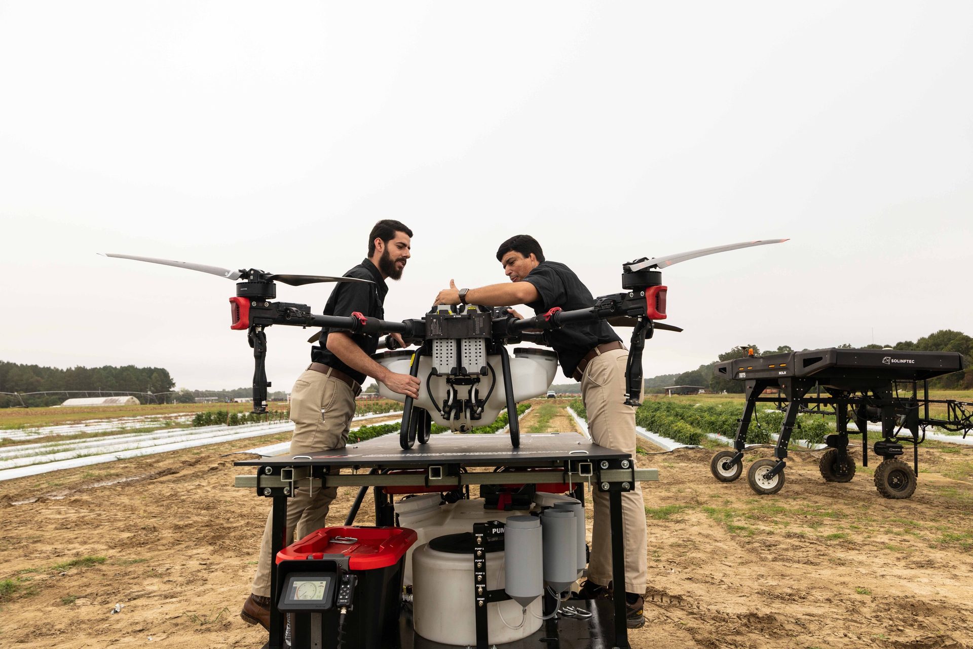 Two men adjust a large agricultural drone on a table in a field, with another robot in the background.