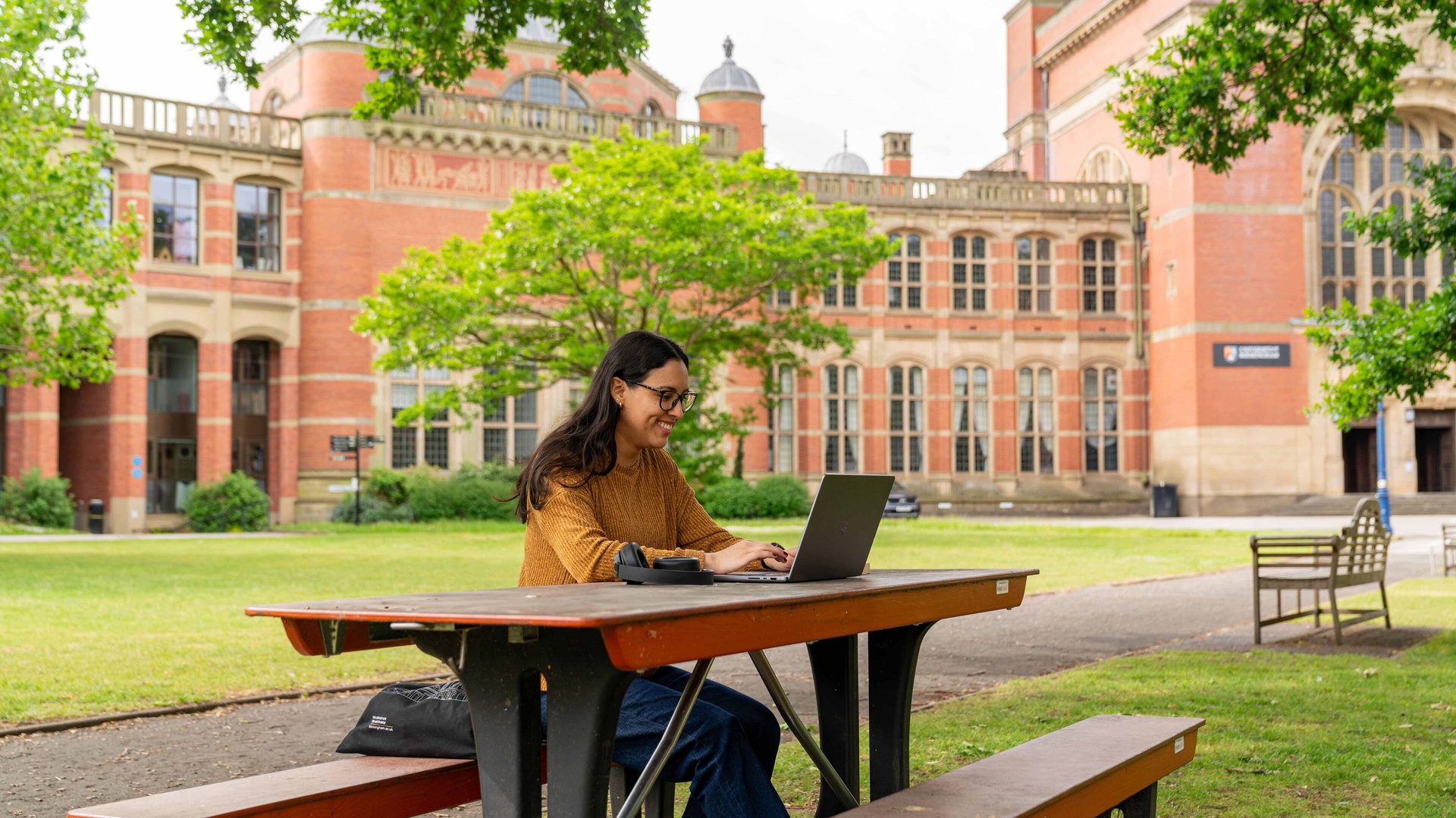 Smiling woman on laptop at outdoor table, grand brick university building behind.