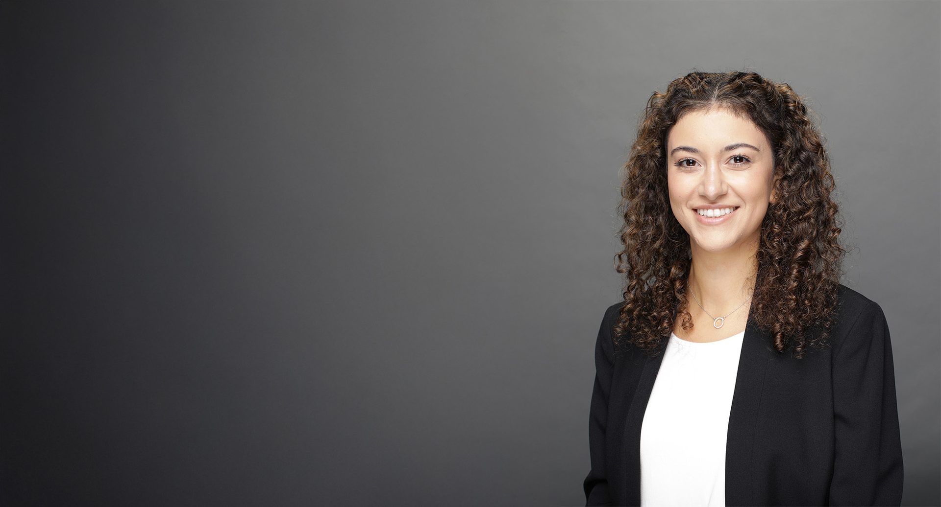 Smiling woman with curly hair in black blazer against a grey background.