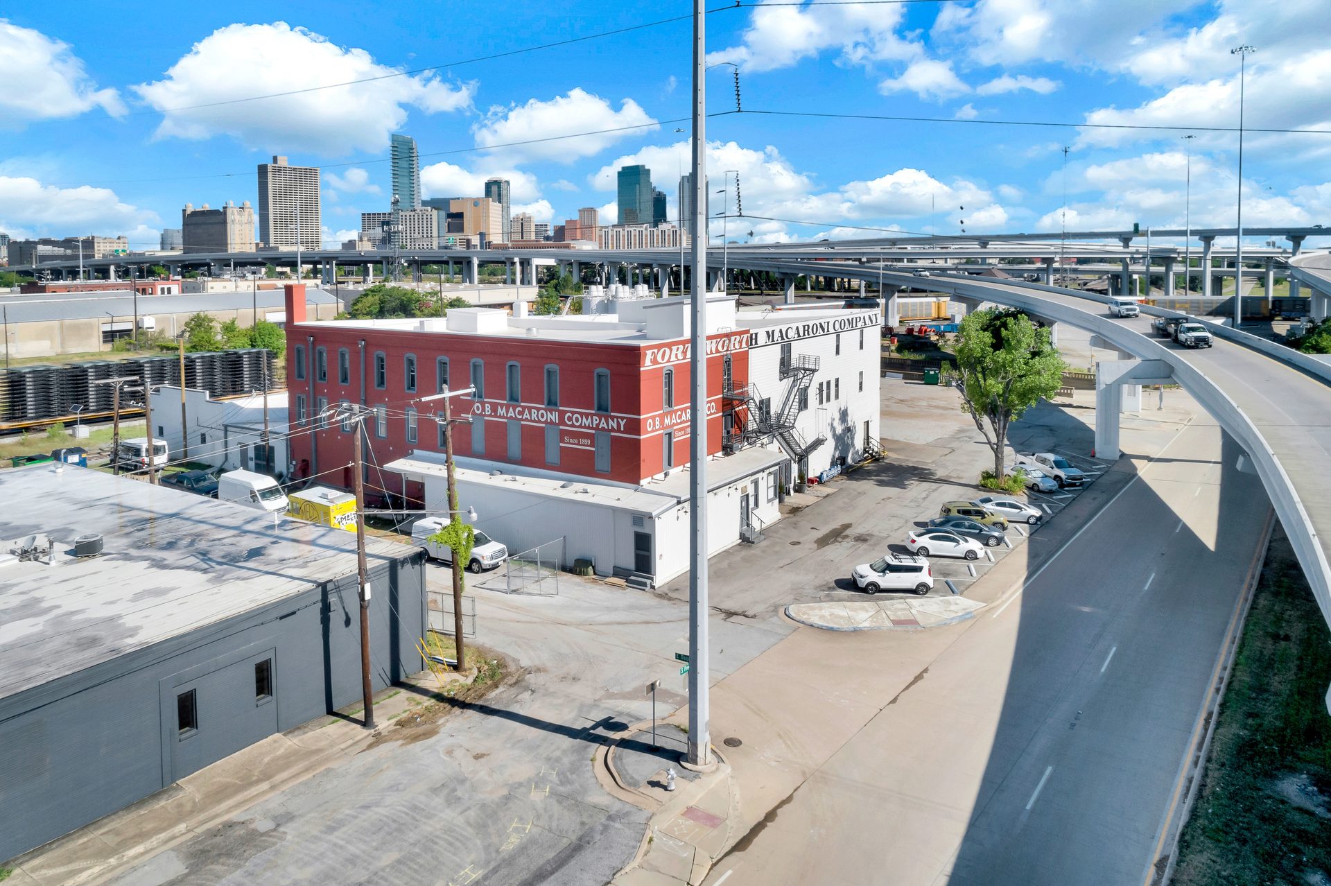 Aerial view of Fort Worth Macaroni Company building, elevated highways, and city skyline under a blue sky.