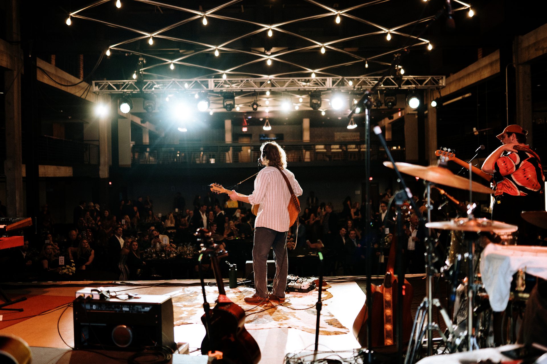 A guitarist performs on a brightly lit stage, seen from behind, with an audience watching.
