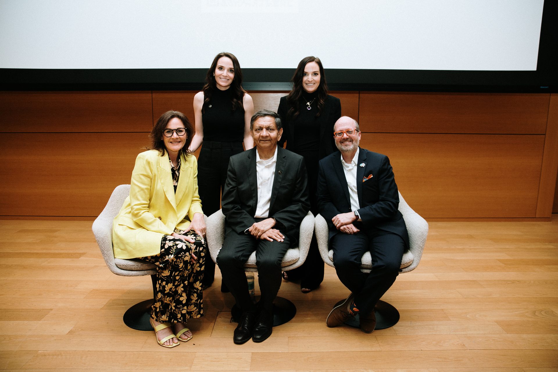 Five people, three seated and two standing, smile for a photo indoors with a screen behind them.