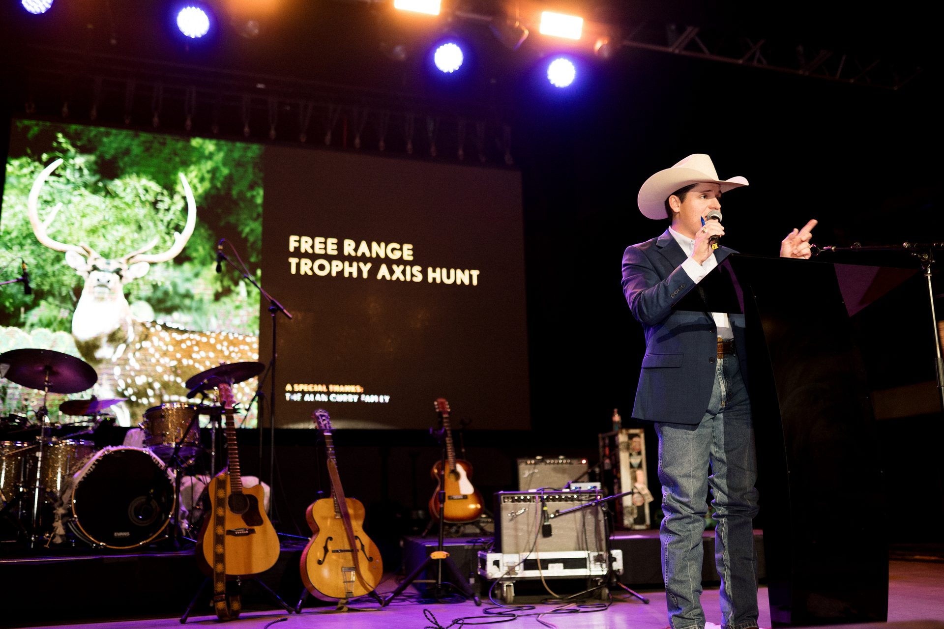 Man in cowboy hat speaks on stage with "FREE RANGE TROPHY AXIS HUNT" screen, deer, and instruments.