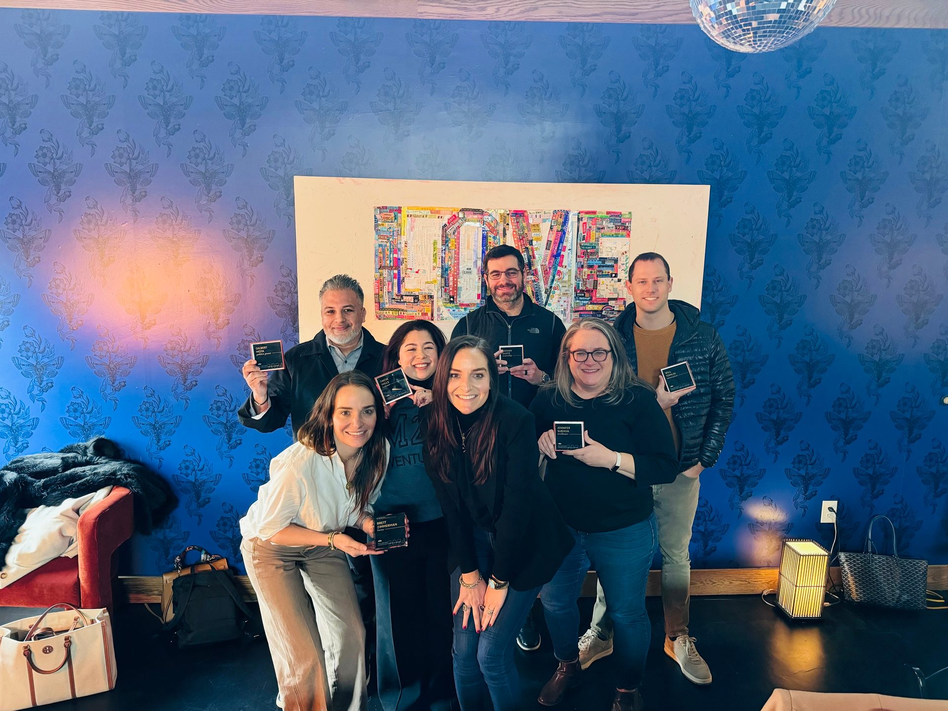 Eight smiling people, mostly holding small boxes, stand in front of a blue patterned wall and a "LOVE" artwork.