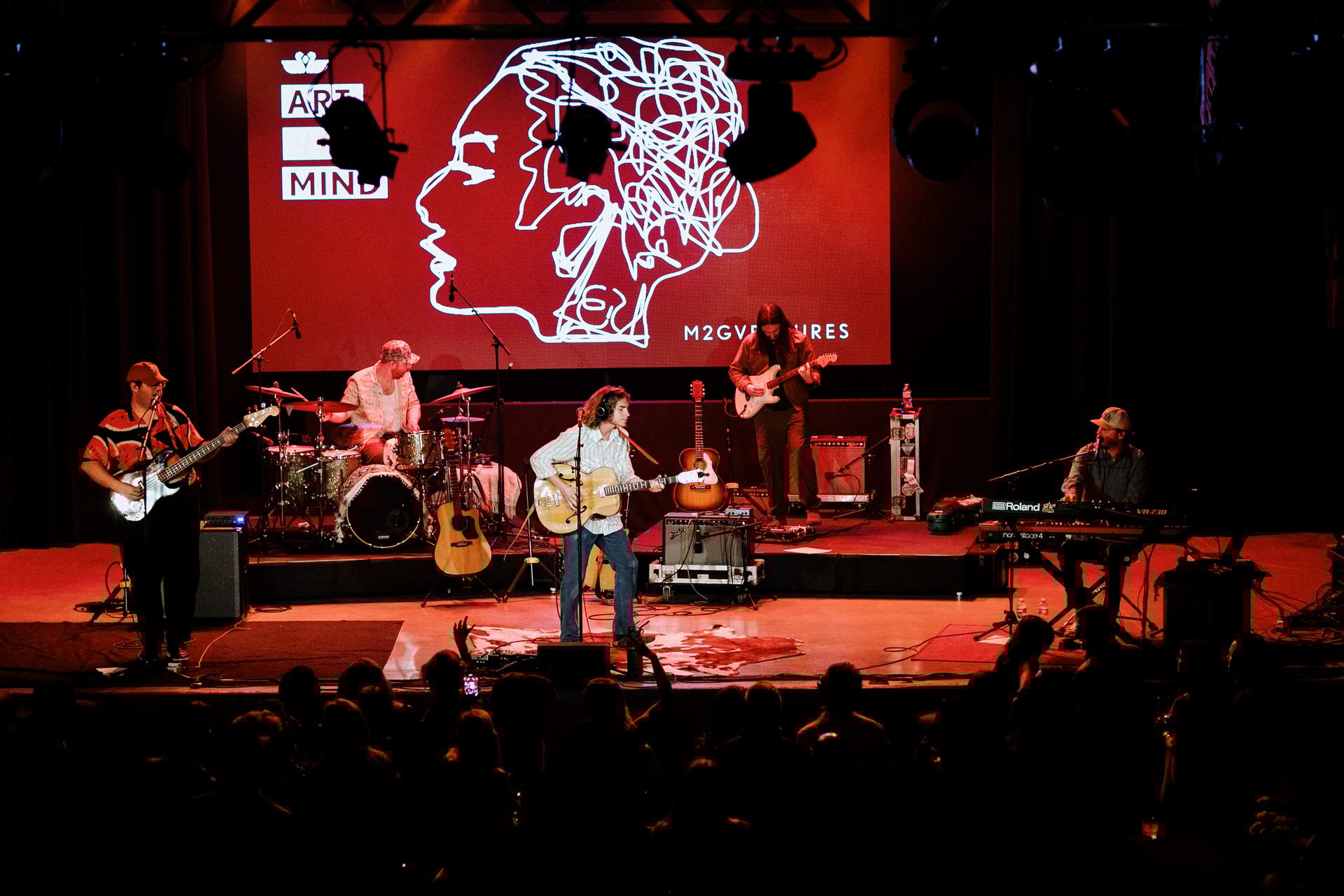 Live band performs on stage with red graphic screen, audience visible.
