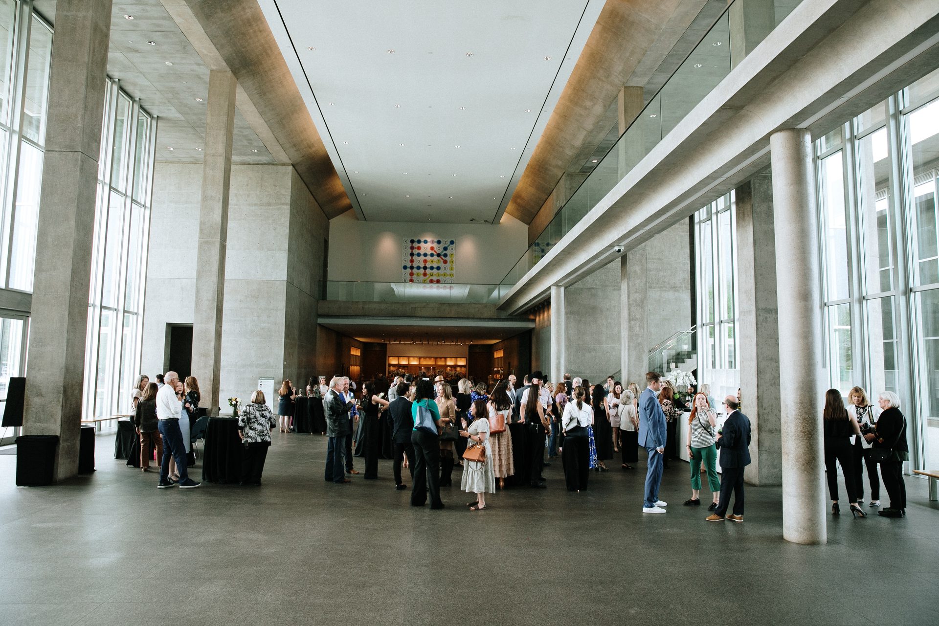 Guests gather in a grand modern lobby with concrete pillars, glass walls, and a colorful artwork.