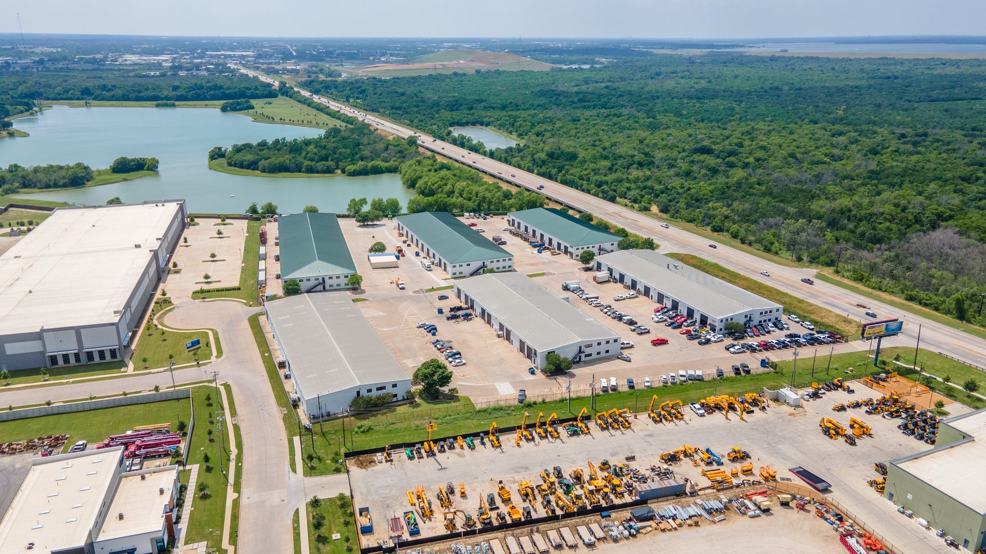 Aerial view of an industrial park next to a lake and highway, featuring warehouses and heavy equipment.
