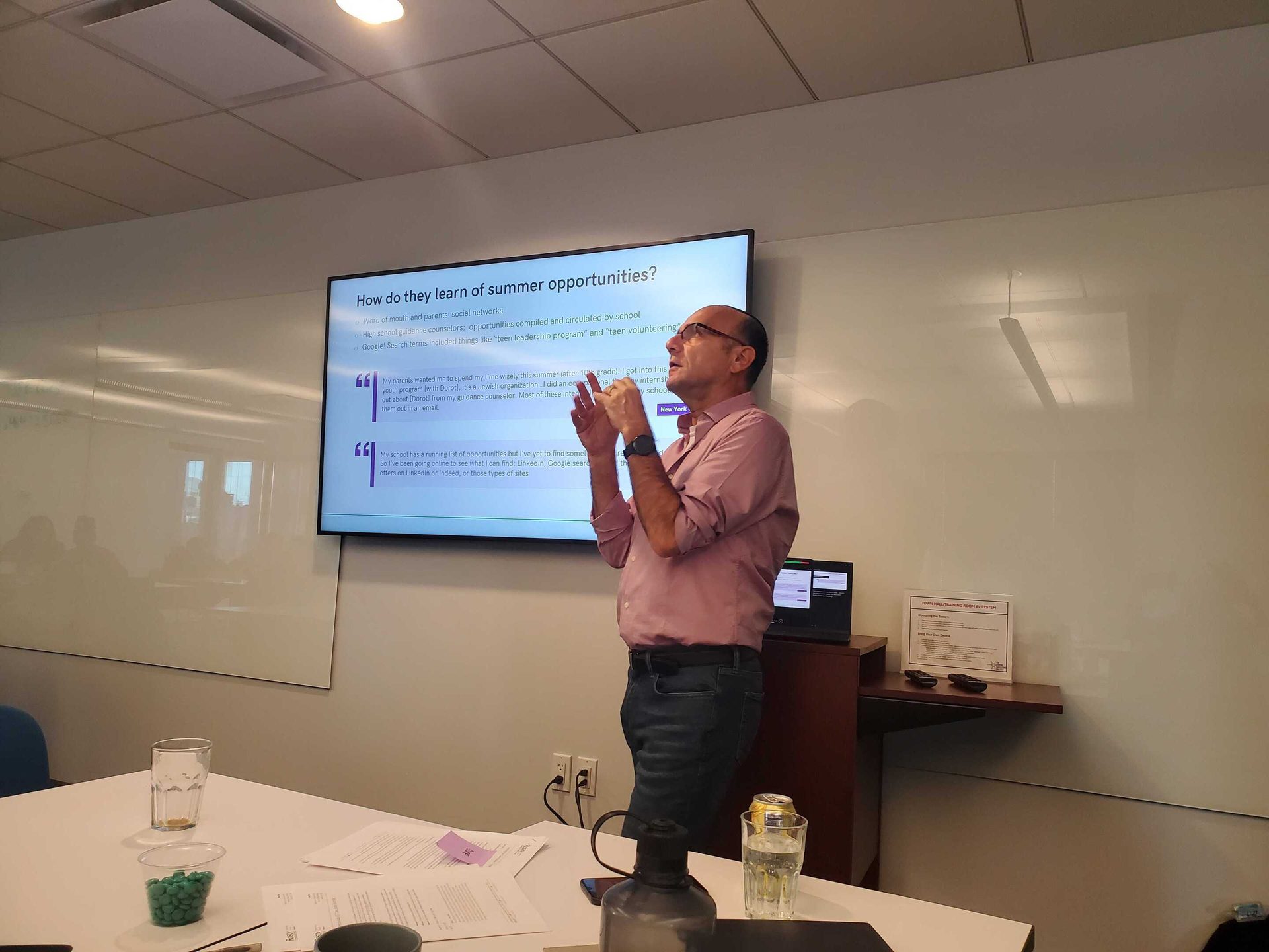 A man presents, gesturing at a screen displaying text about summer opportunities.