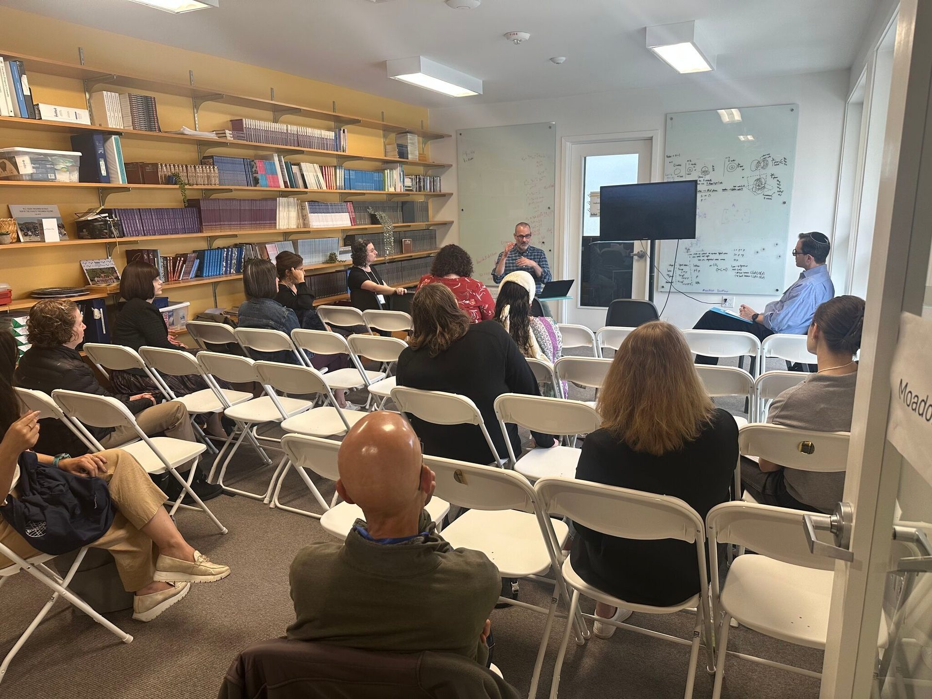 An audience watches a speaker in a room with bookshelves and a whiteboard.