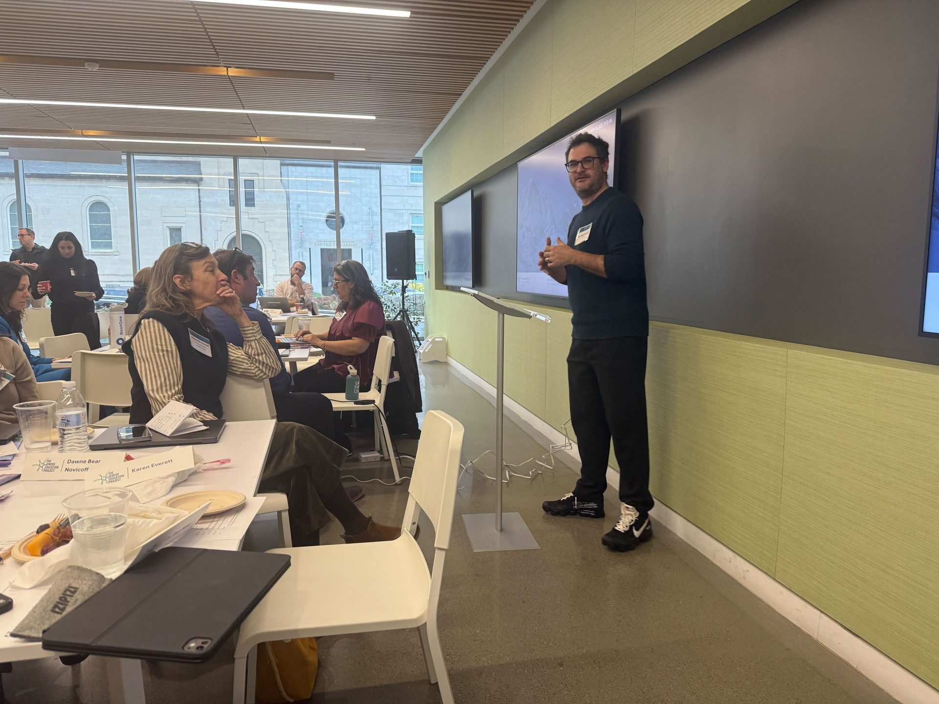 Man presenting to an audience seated at tables in a modern conference room.