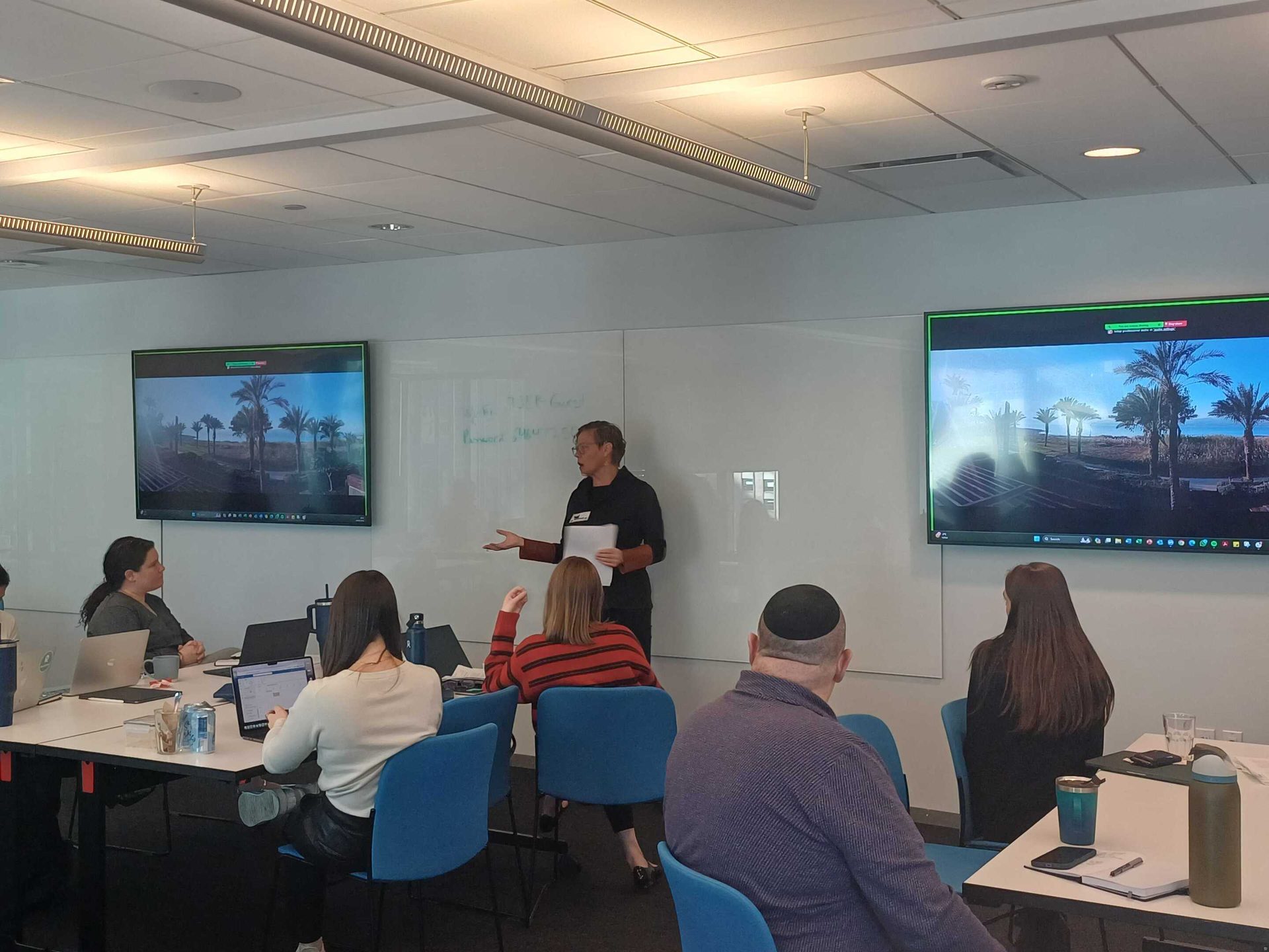 A presenter addresses an audience in a meeting room with large screens displaying palm trees.