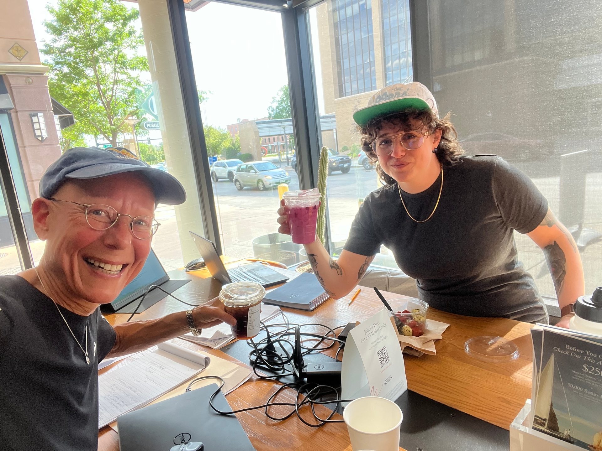 Two people with drinks and laptops at a cafe table by a window, one smiling at the camera.