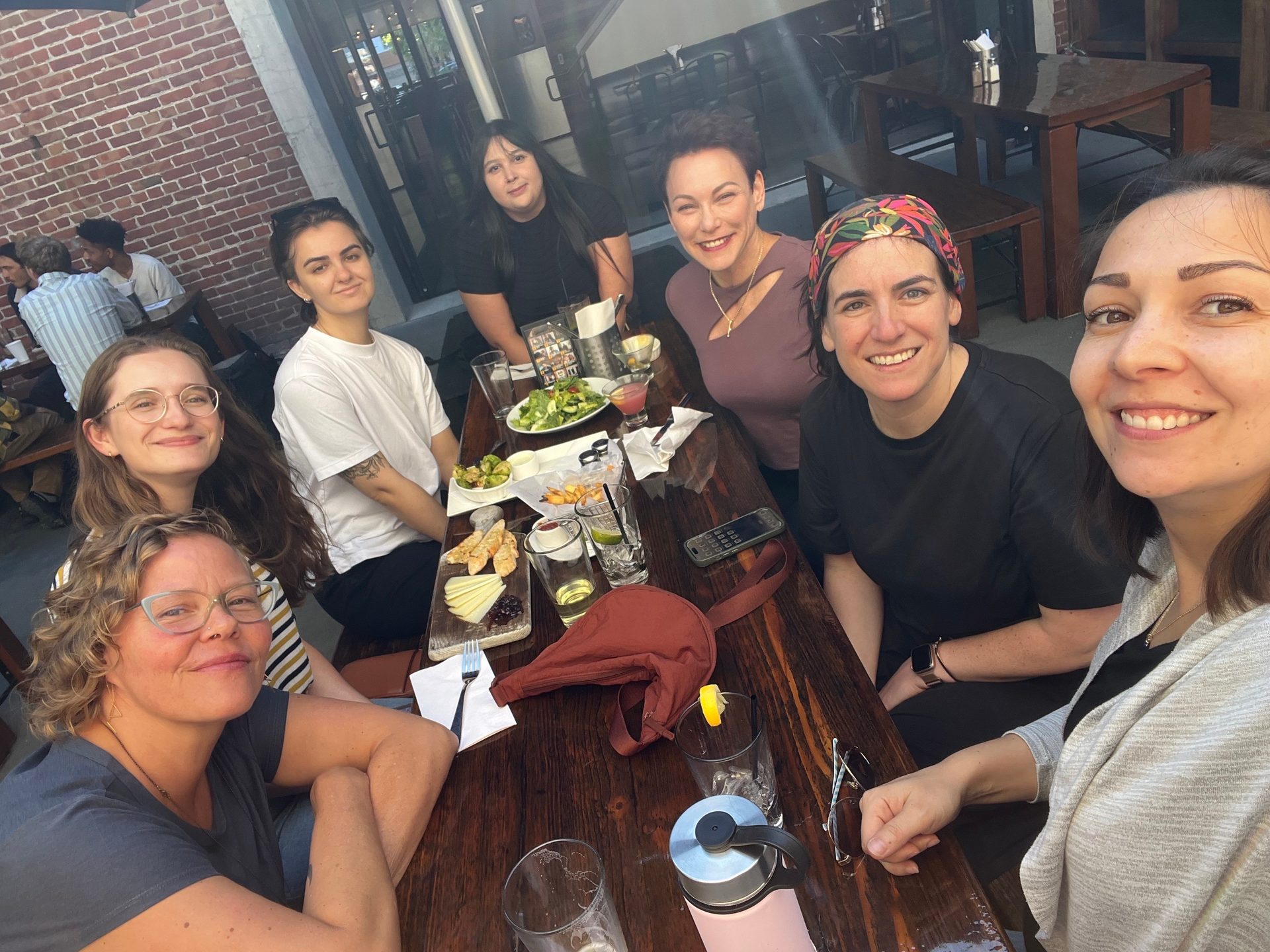 A group of smiling women sitting around a table with food and drinks at an outdoor restaurant.