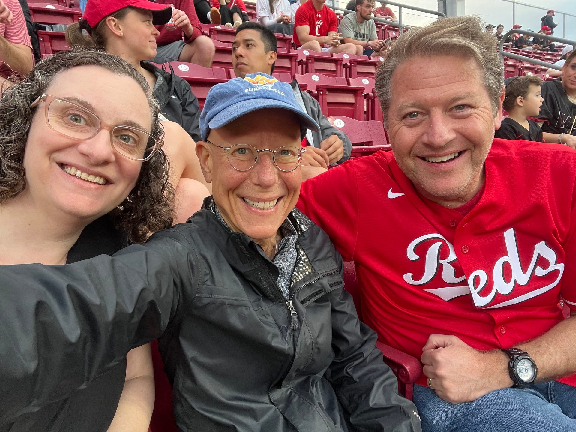 Three smiling fans, one in a 'Reds' jersey, taking a selfie at a stadium.