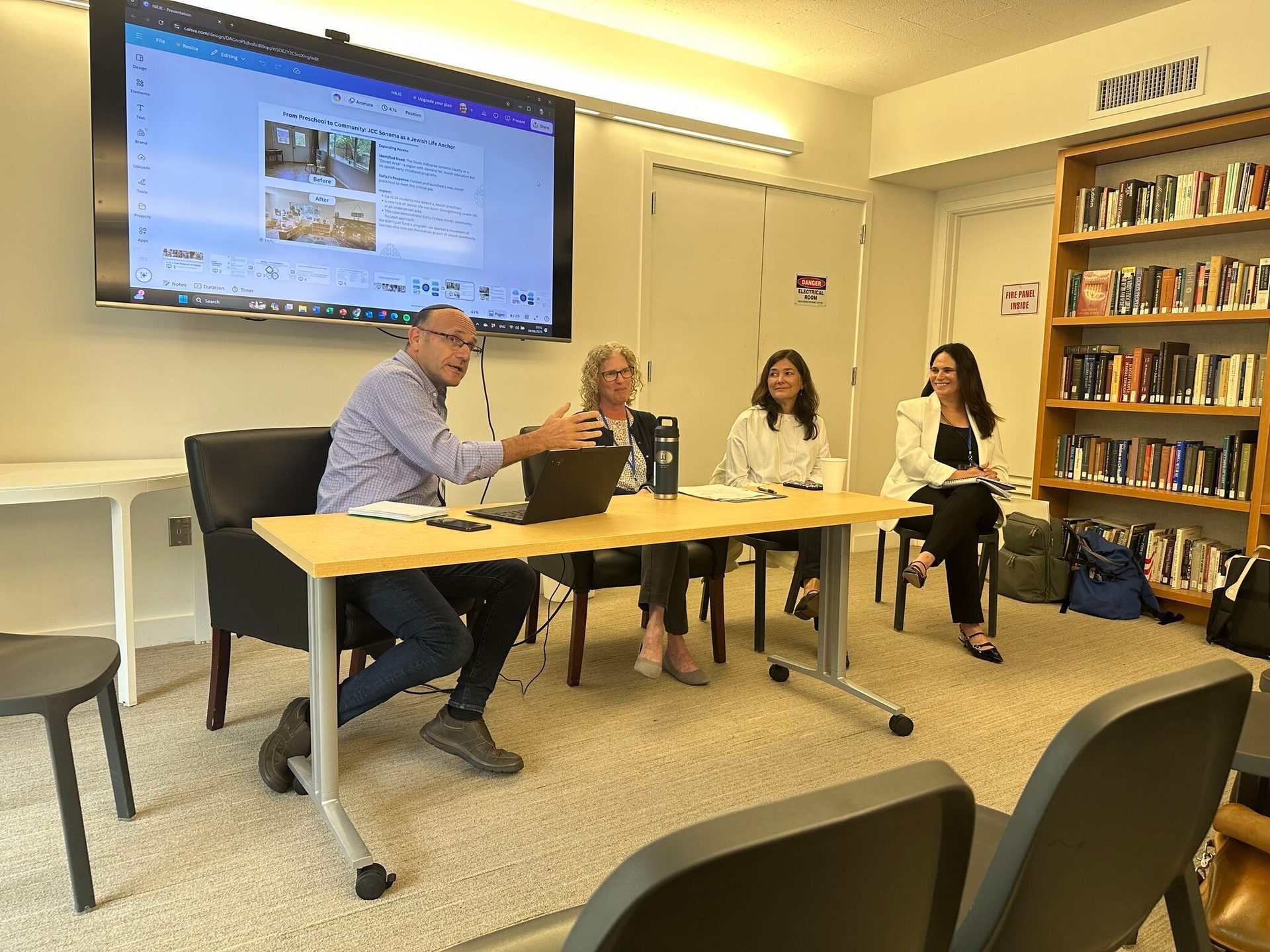 A man speaks to three women at a table, in front of a presentation screen and bookshelves.
