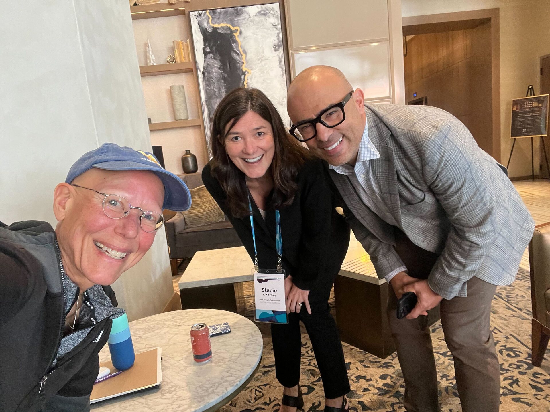 Three smiling people: one in a blue cap, one with an ID badge, and a man in glasses and a suit jacket.
