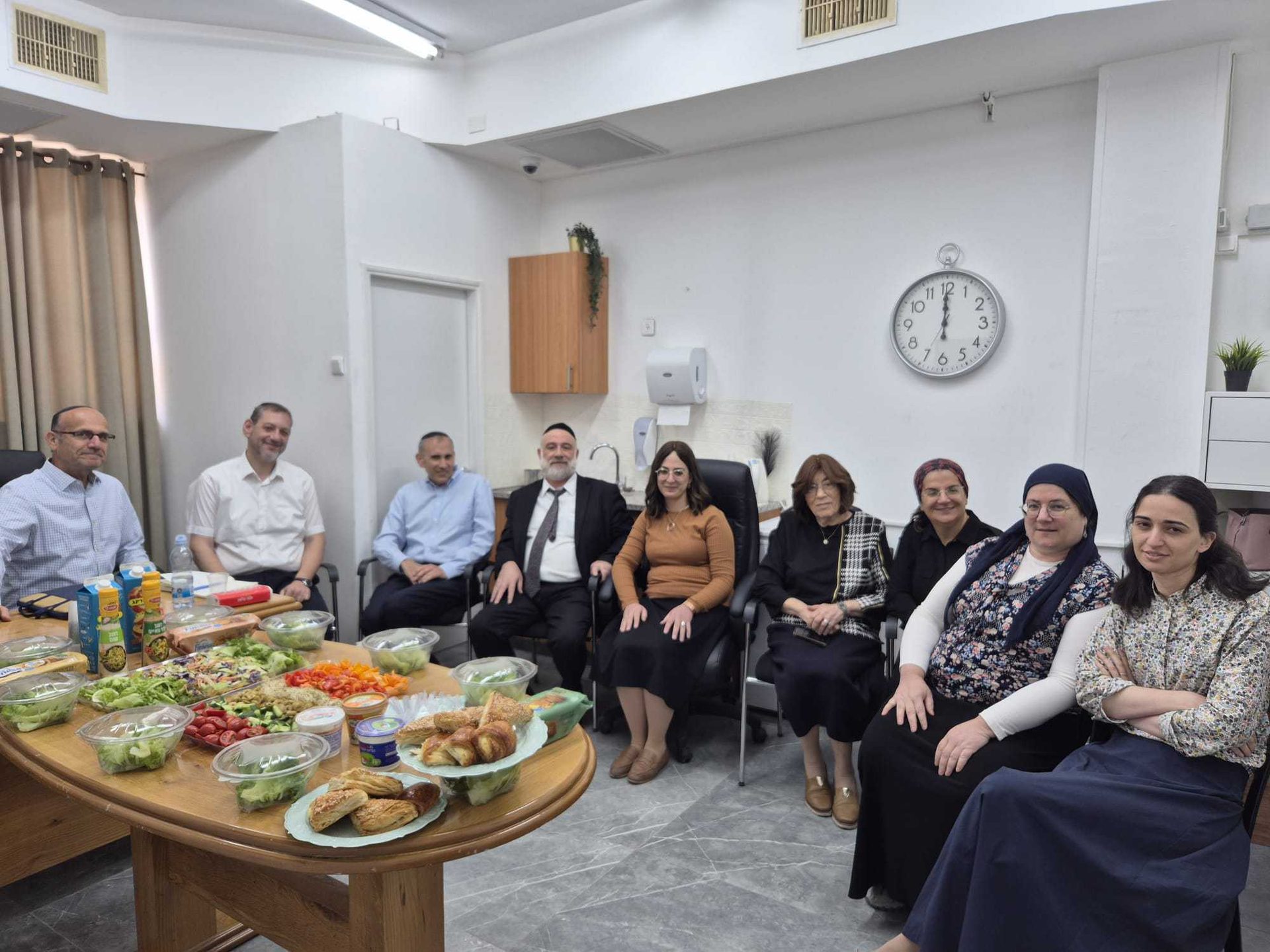 A group of men and women sharing a meal of salads and pastries around a table indoors.
