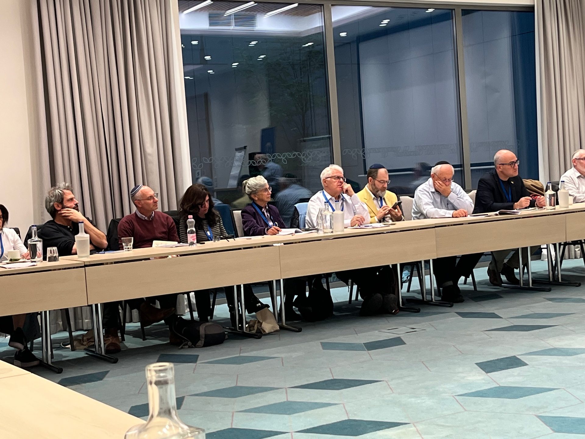 Attendees, some wearing kippahs, sit at a long table during a meeting or conference.
