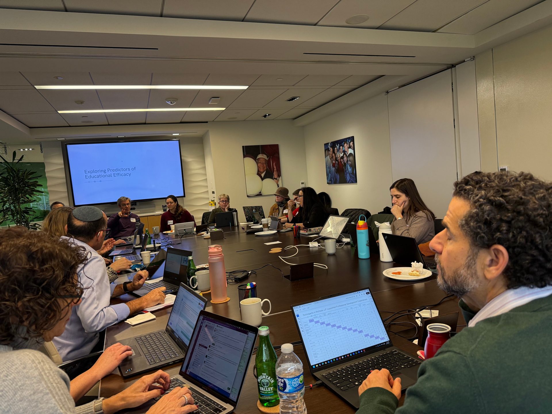 People in a meeting room, working on laptops around a large table, with a presentation screen.