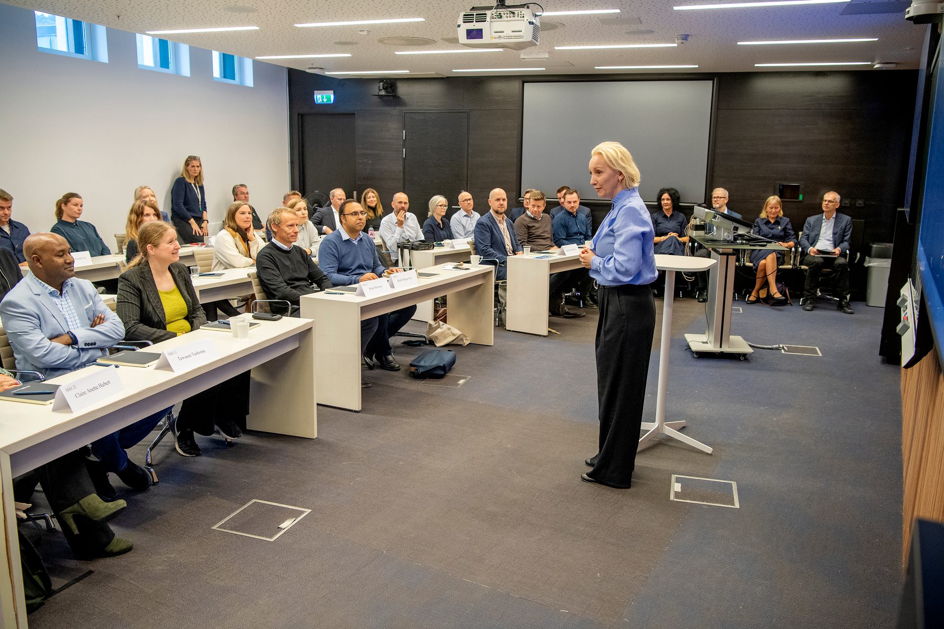 A blonde woman presents to a diverse group of professionals seated at tables in a modern meeting room.