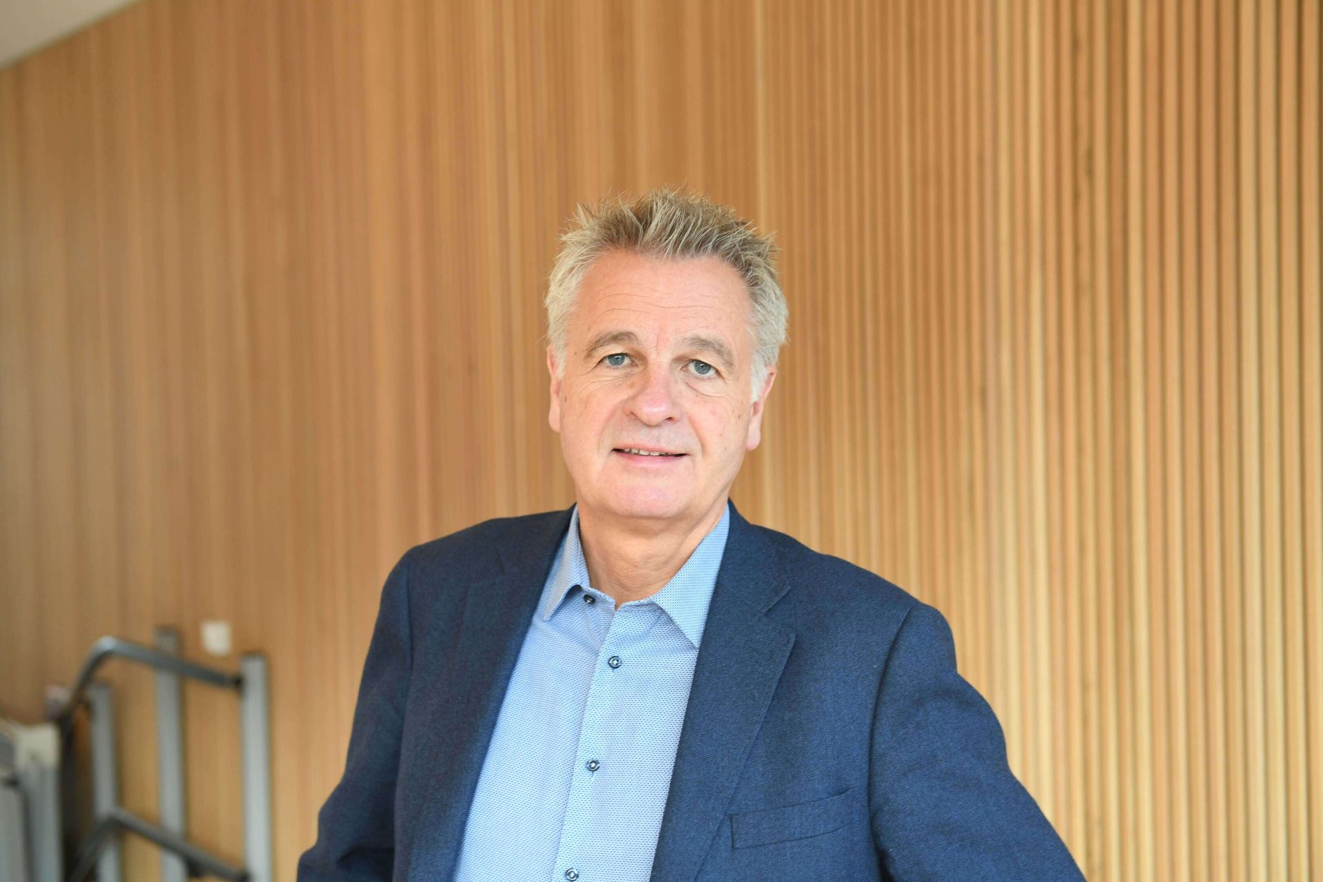 A man in a blue suit and light blue shirt smiles at the camera, standing against a wood-paneled wall.