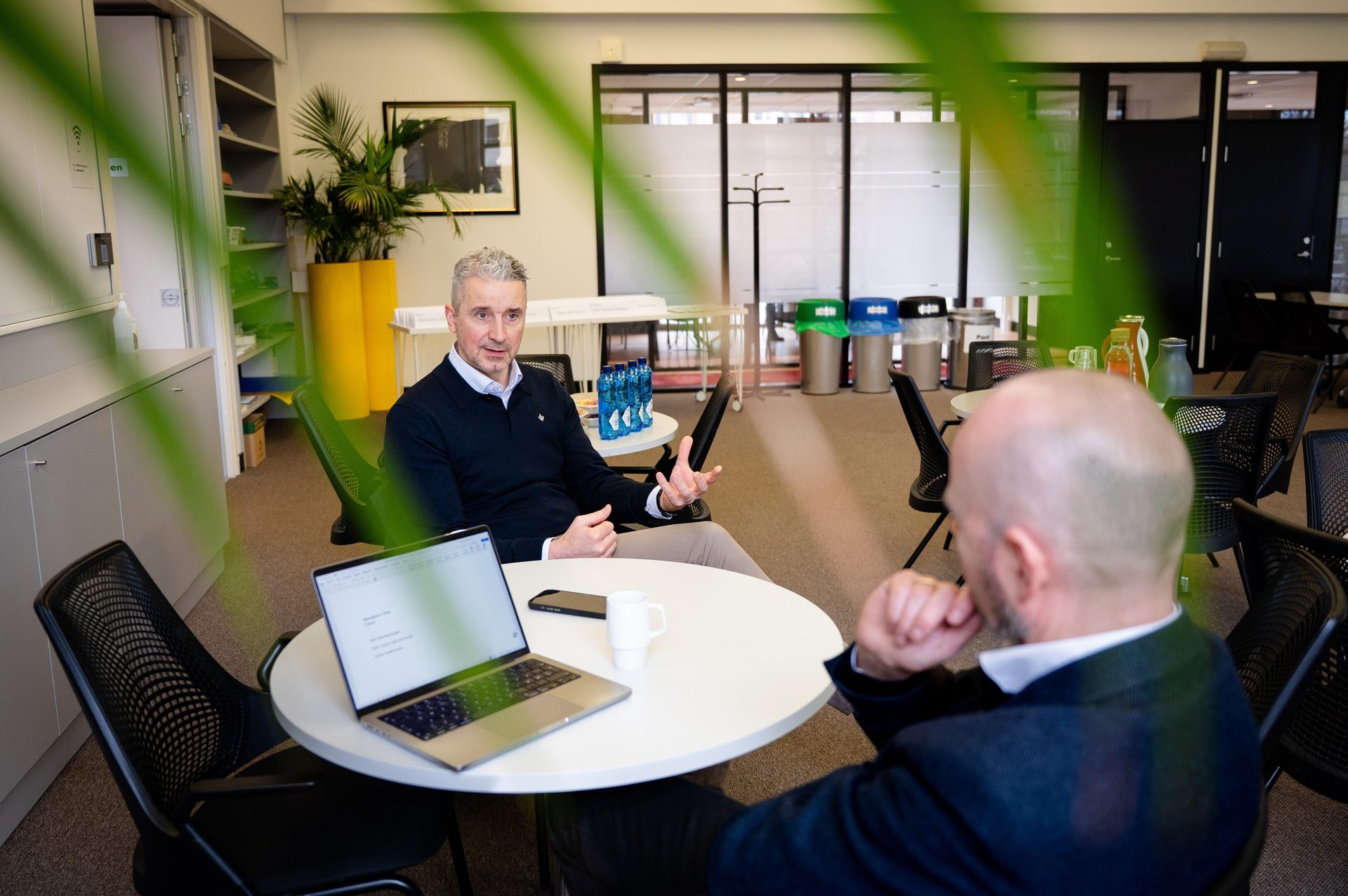 Two men discuss around a table with a laptop in a modern office setting.