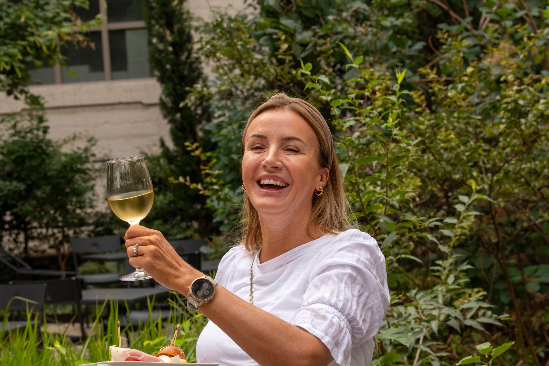 A laughing woman holding a glass of white wine and appetizers outdoors.