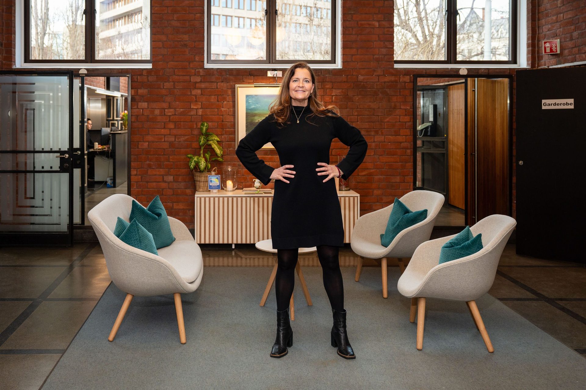 A smiling woman in a black dress stands confidently with hands on hips in a modern office lobby.