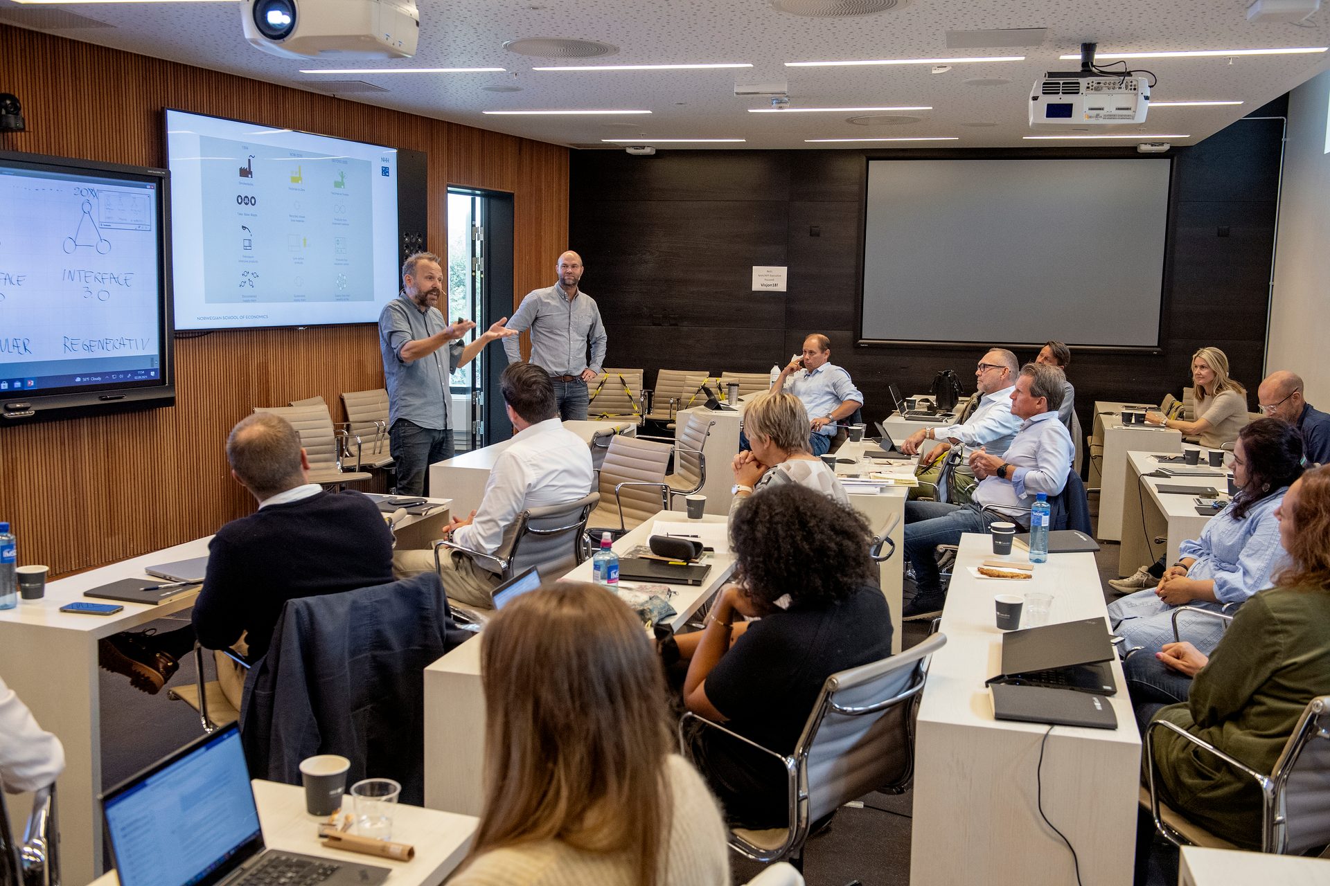 Attendees listen to two presenters in a modern conference room with large screens displaying information.
