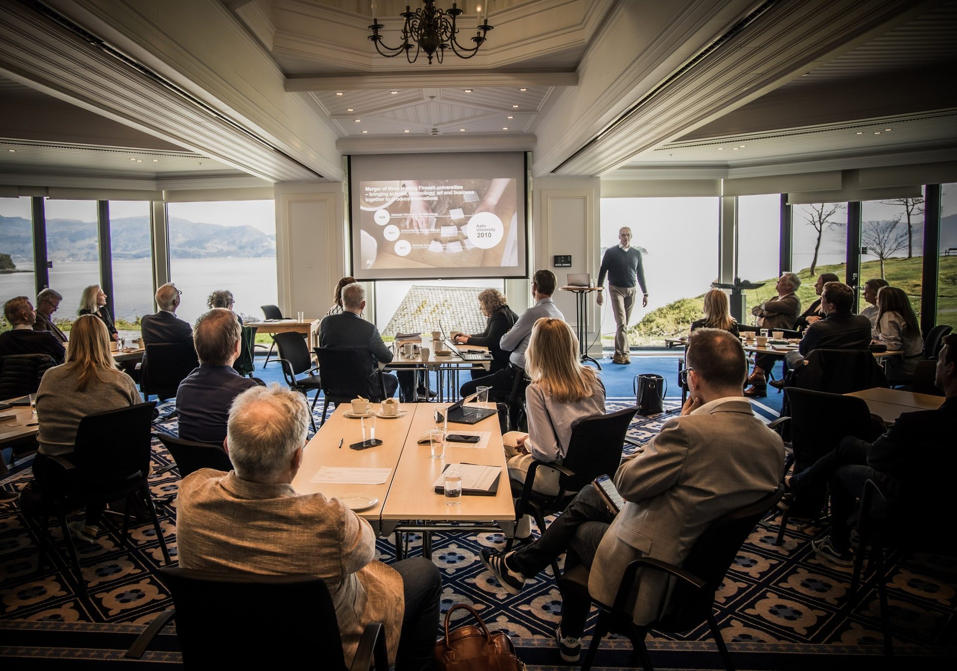 Attendees watch a presentation on a screen in a conference room with large windows overlooking a scenic view.