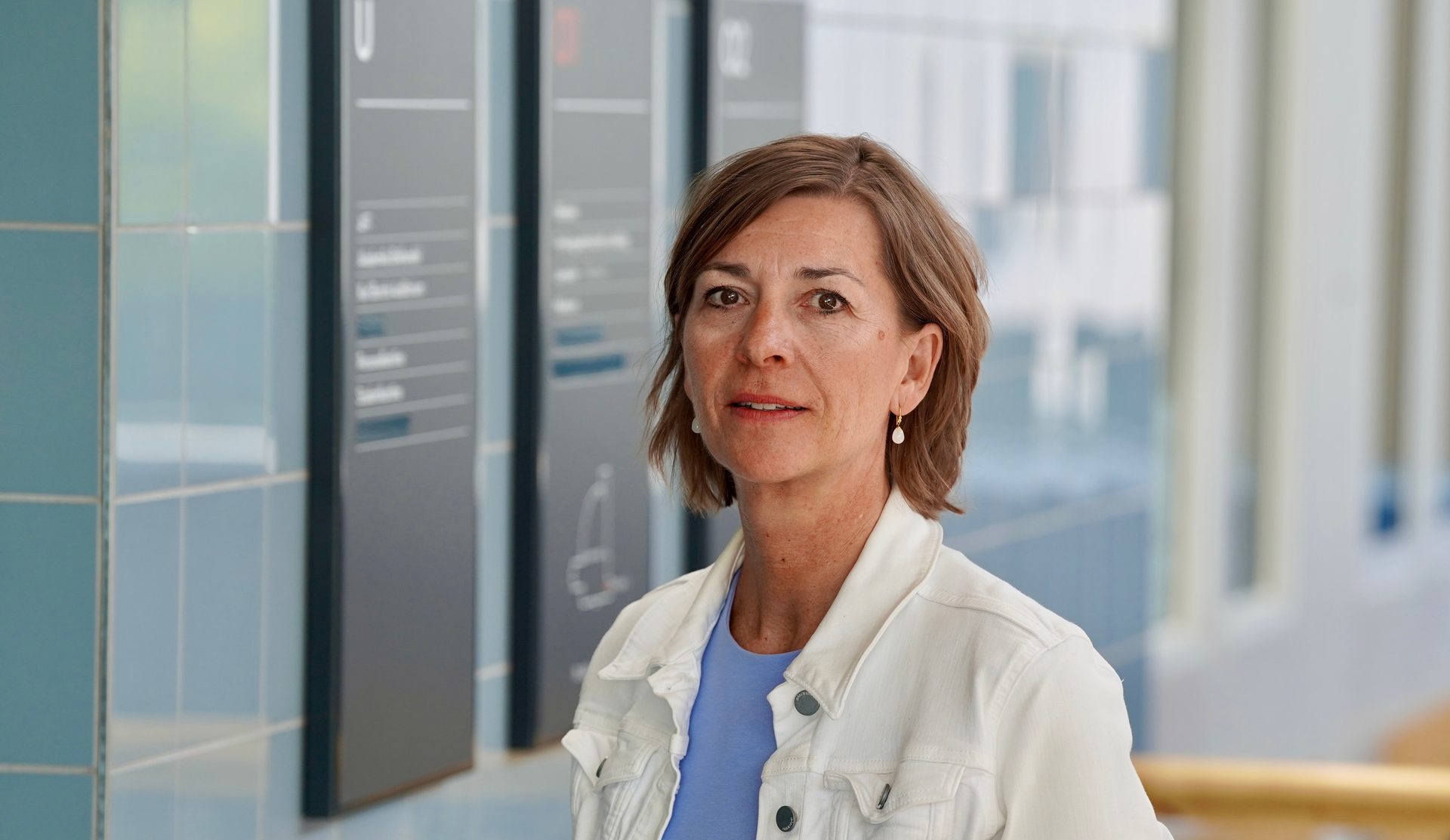 A woman in a white jacket and blue top looks at the camera in an indoor setting.