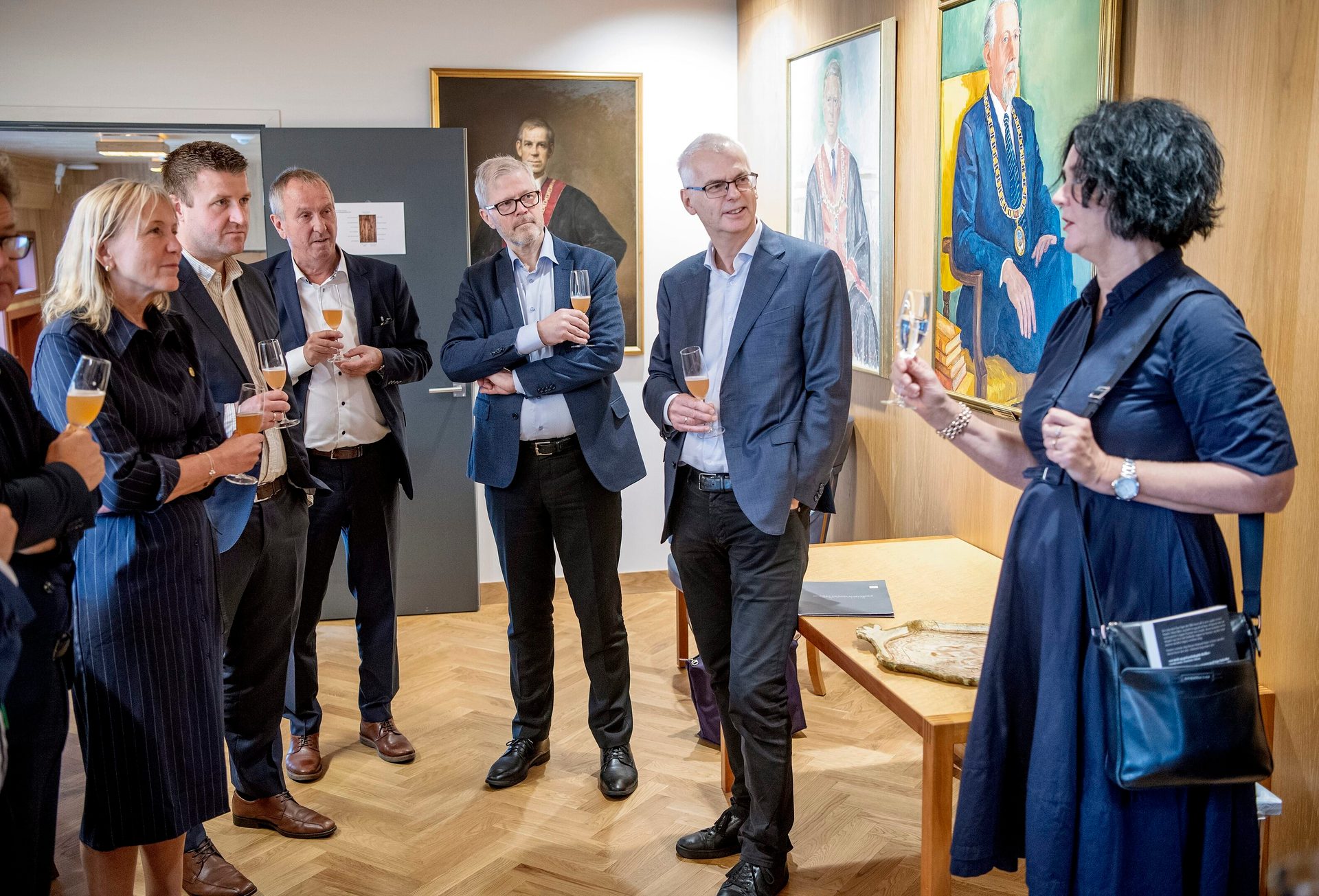 A group of people, mostly men, holding champagne glasses in a room with paintings, a woman speaking.