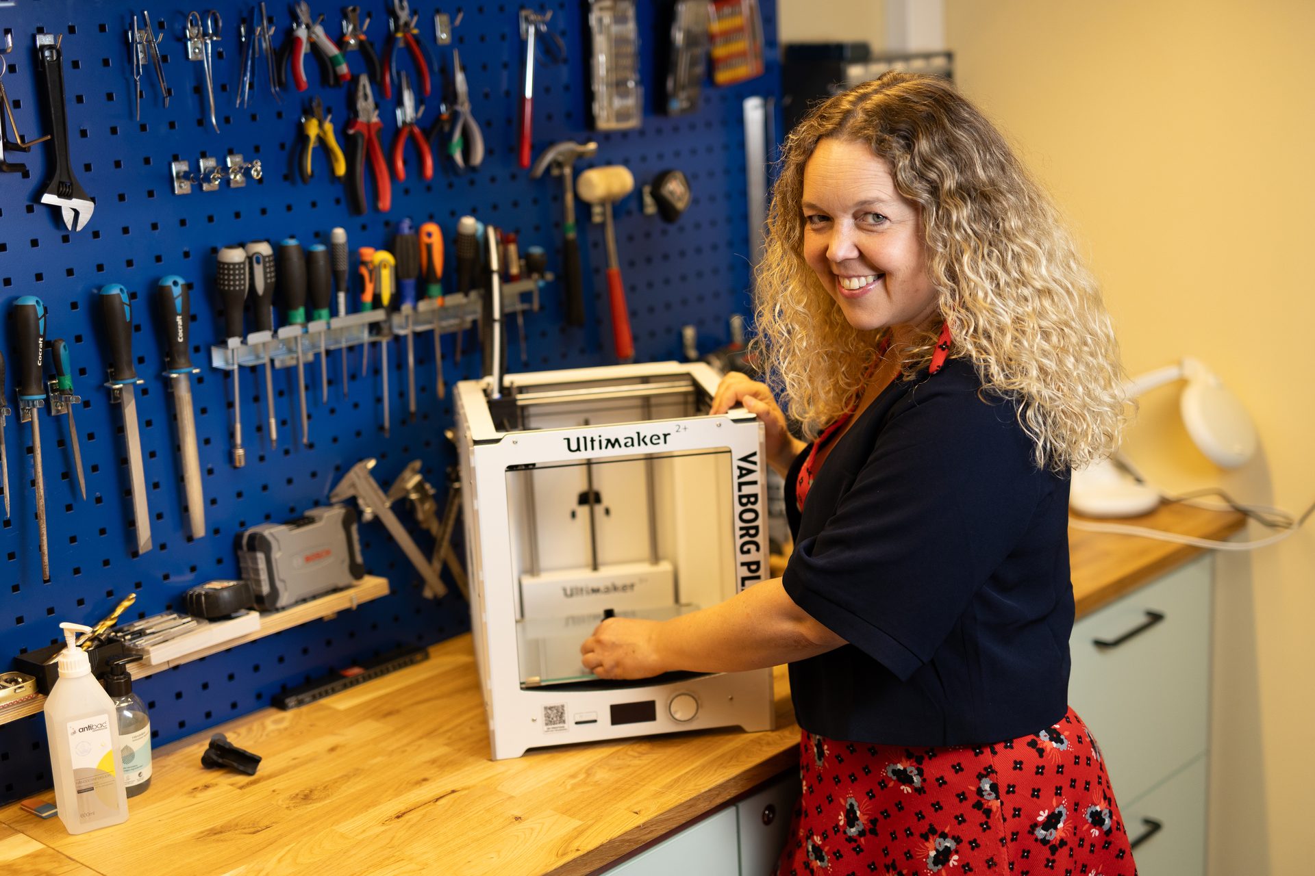 A smiling woman with curly hair operates a white 3D printer on a wooden workbench, with tools on a blue pegboard.